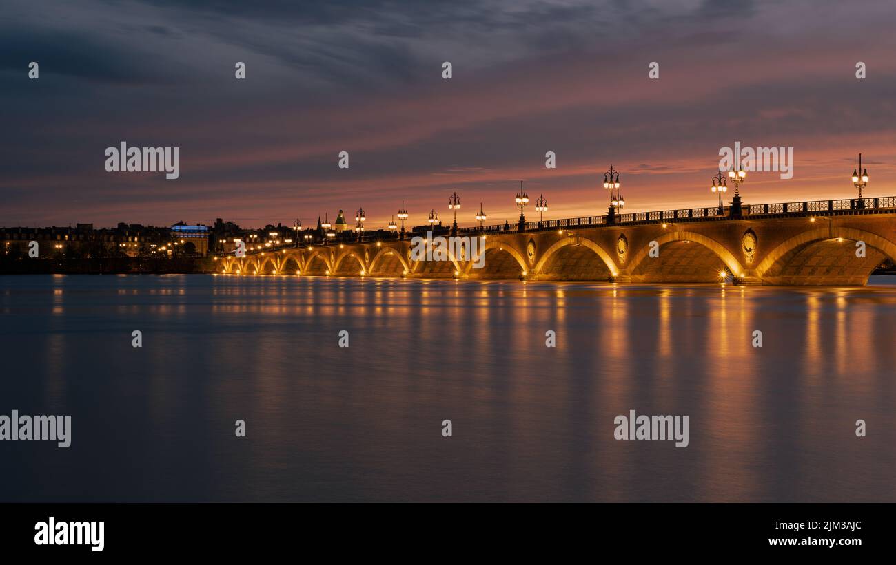 Pont de Pierre, der den Fluss Garonne in der Stadt Bordeaux überspannt, beleuchtet bei Sonnenuntergang mit der Basilique Saint-Michel im Hintergrund Stockfoto