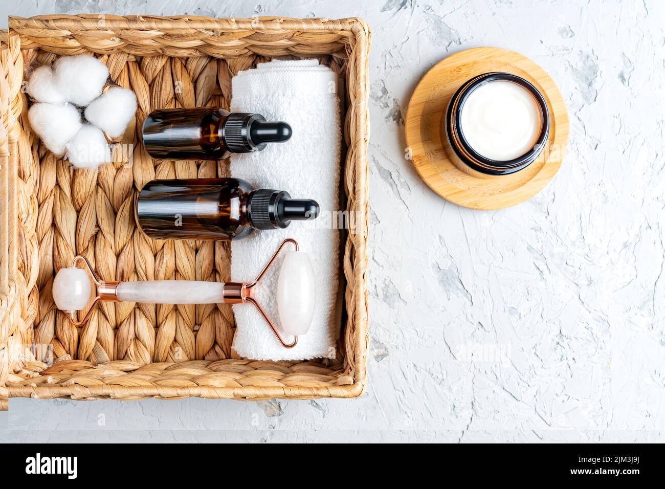 Draufsicht auf Flaschen aus dunklem Glas ohne Marken mit Hautpflegeprodukten in einem Öko-Korb. Gesicht Massage Roller und Baumwollblume. Gesichtsbehandlung Set für Home p Stockfoto