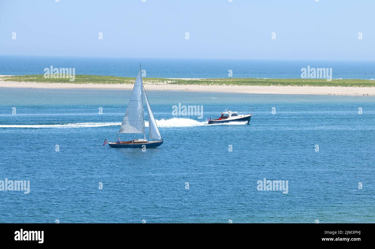Ein Motorboot, das an einem Segelboot im Kanal vor Lighthouse Beach, Chatham, Massachusetts, auf Cape Cod, USA, vorbeifährt. Chatham Bar im Hintergrund Stockfoto