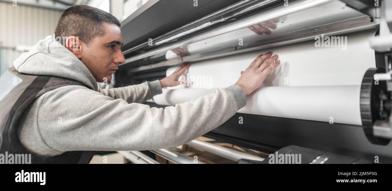 Der Bediener des Technikers wechselt die Papierrolle auf einem großen hochwertigen Industriedrucker und Plottergerät im digitalen Druckereibüro Stockfoto