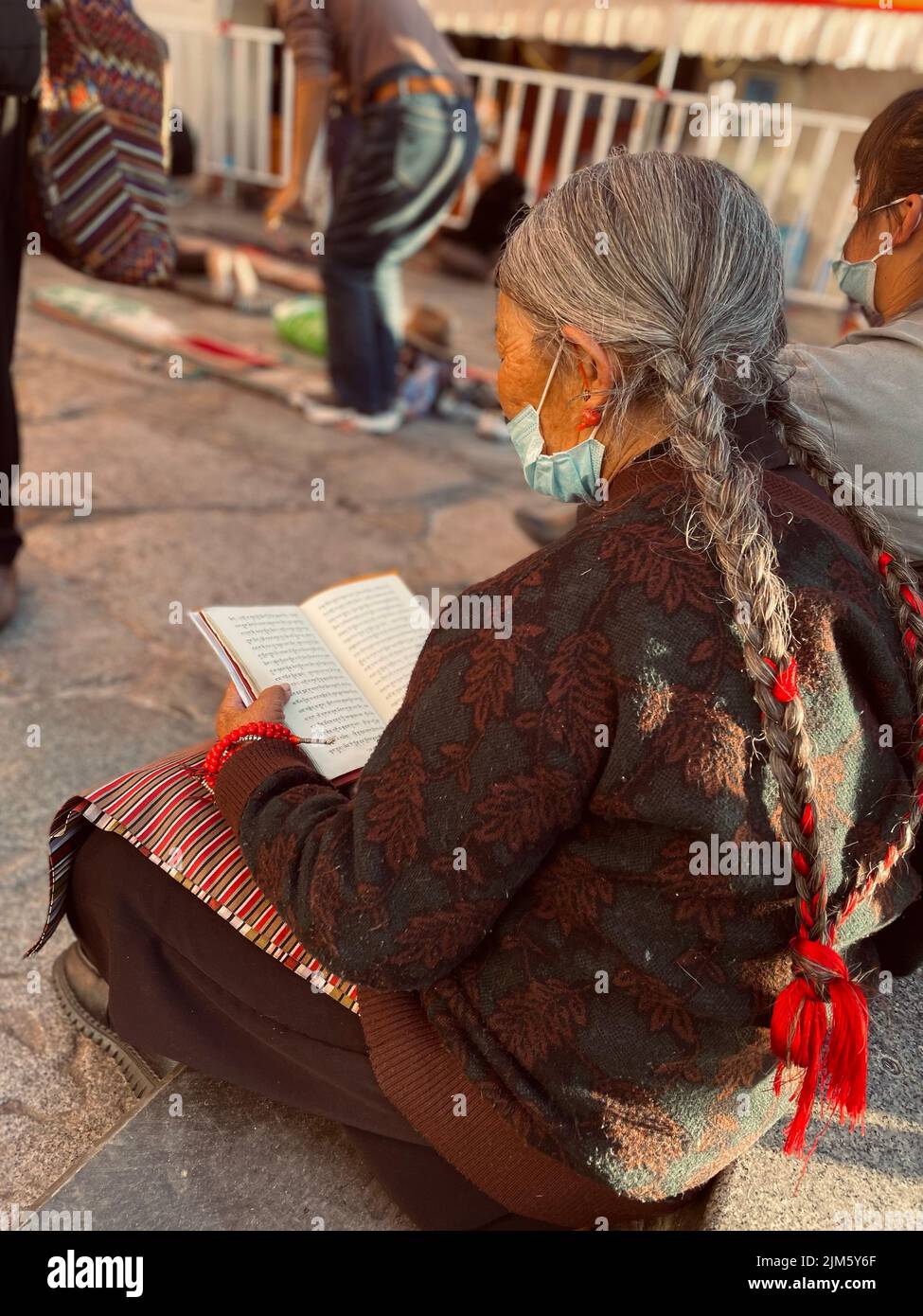 Eine Rückaufnahme einer alten Frau mit Zöpfen, die auf dem Boden sitzen und ein Buch in Tibet, China, lesen Stockfoto