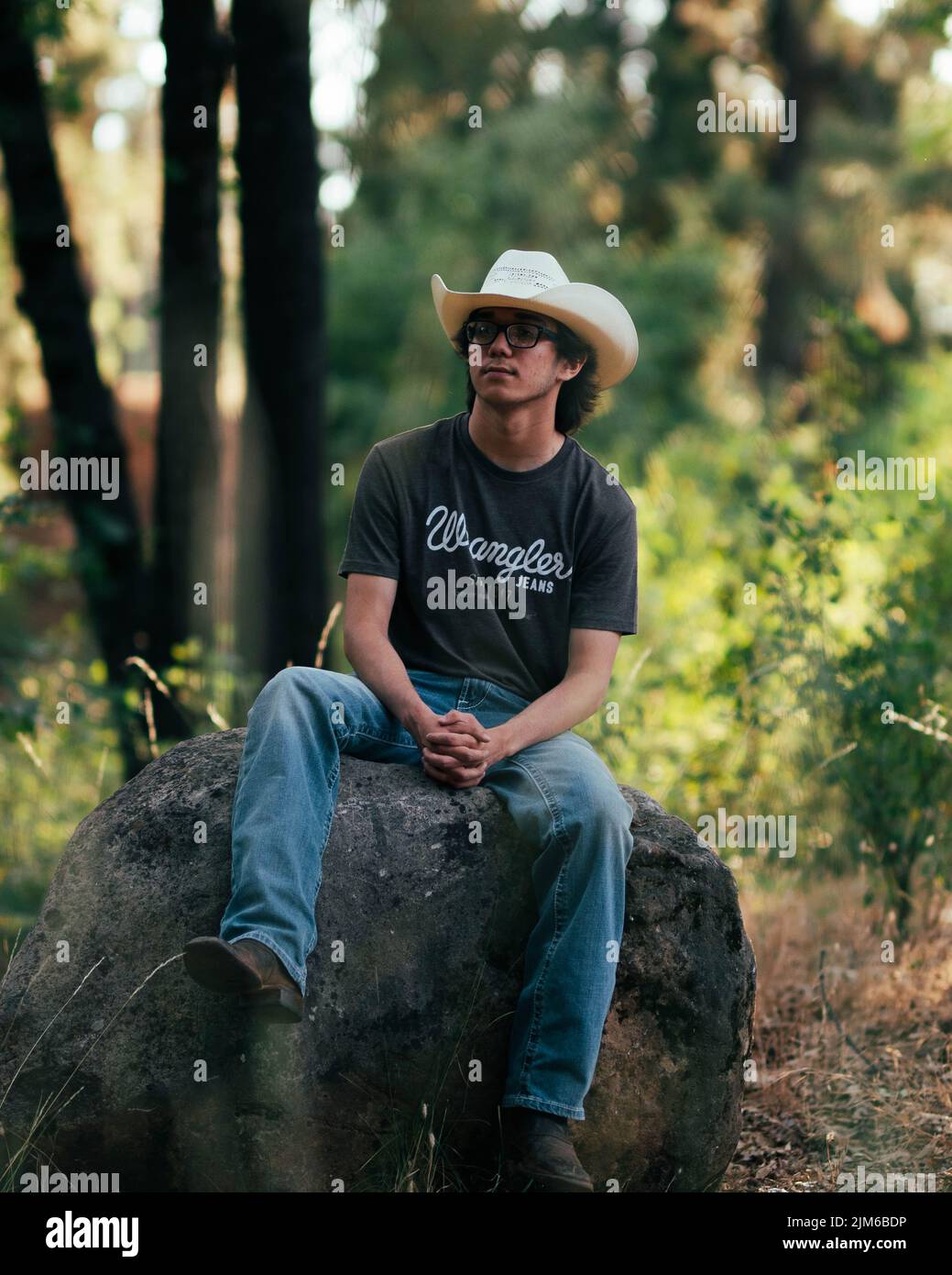 Ein Mann mit einem weißen Cowboyhut und einer Brille, der auf einem Felsen sitzt, mit Blick auf die Natur im Hintergrund Stockfoto