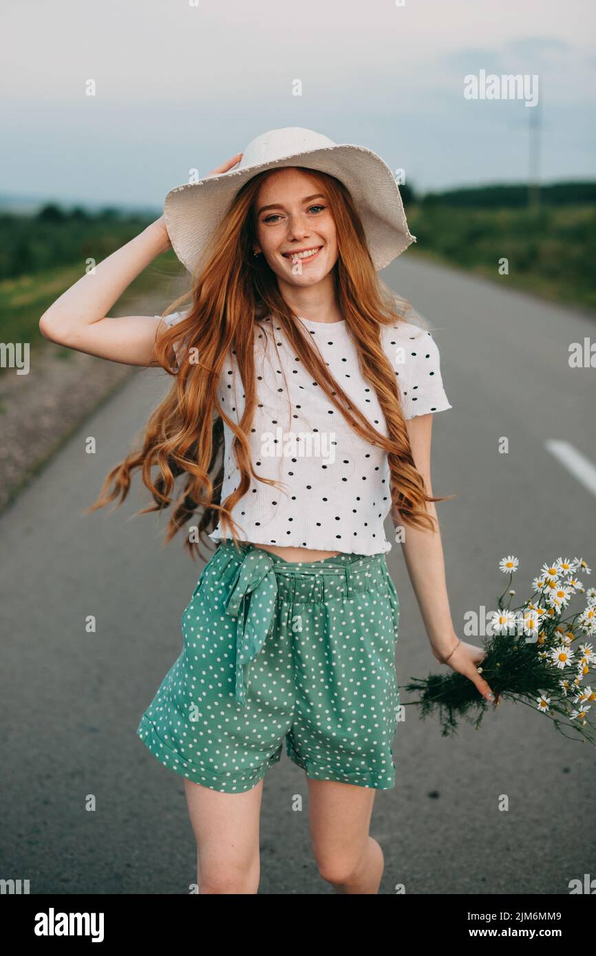 Reizende rothaarige Mädchen in Sonnenhut, halten schöne Blumen lächeln an der Kamera, während Sie auf der Landstraße zu Fuß. Elegantes Modell posieren in der Natur. Stockfoto