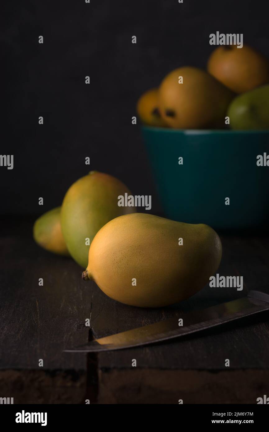 Reife Mangos, tropische Früchte auf der Tischplatte, mit Obstschale und Messer, dunkler Hintergrund mit Weichfokus, Kopierraum Stockfoto