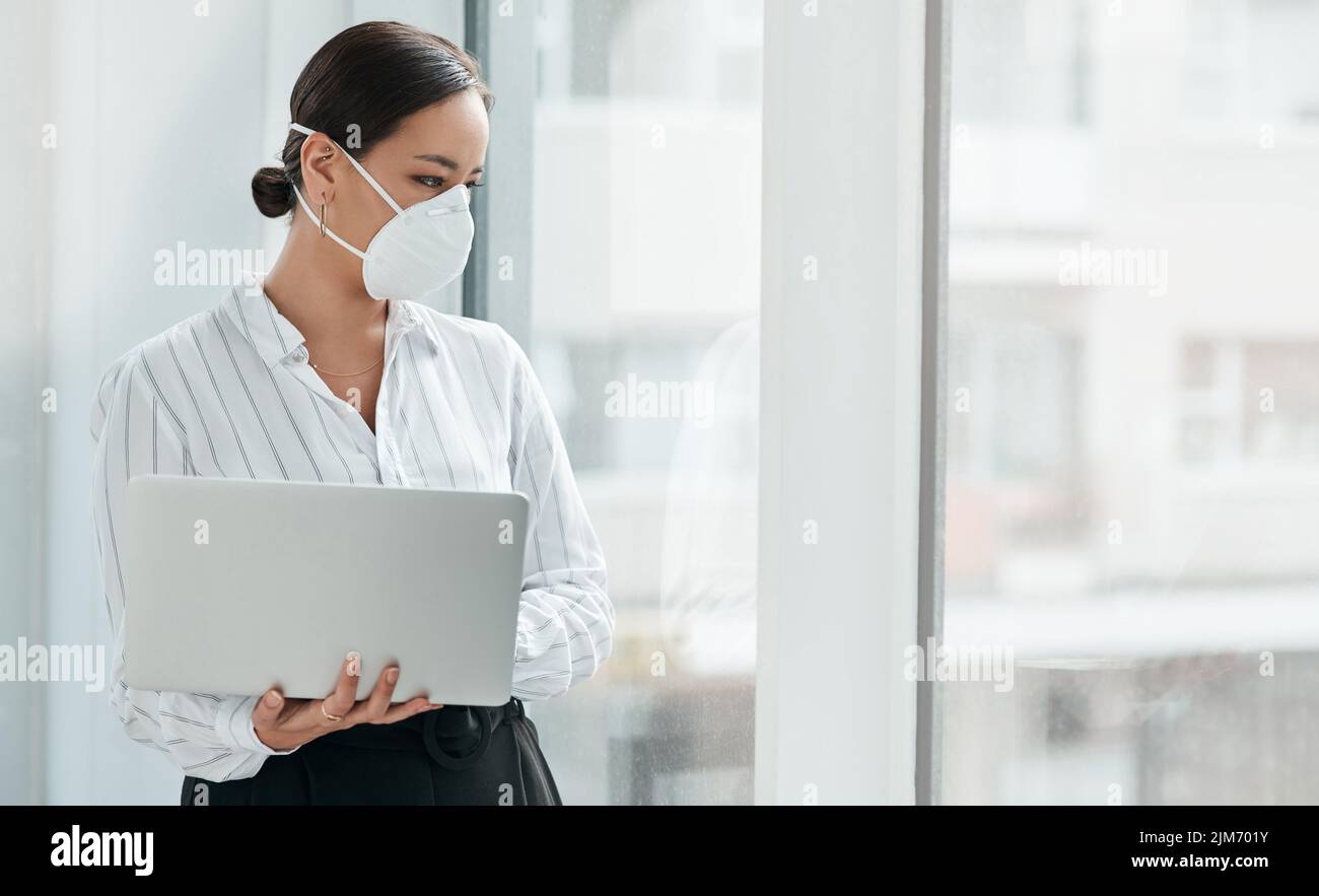 Eine Chance, Dinge anders zu machen: Eine maskierte junge Geschäftsfrau, die einen Laptop in der Hand hält und aus einem Fenster in einem modernen Büro blickt. Stockfoto