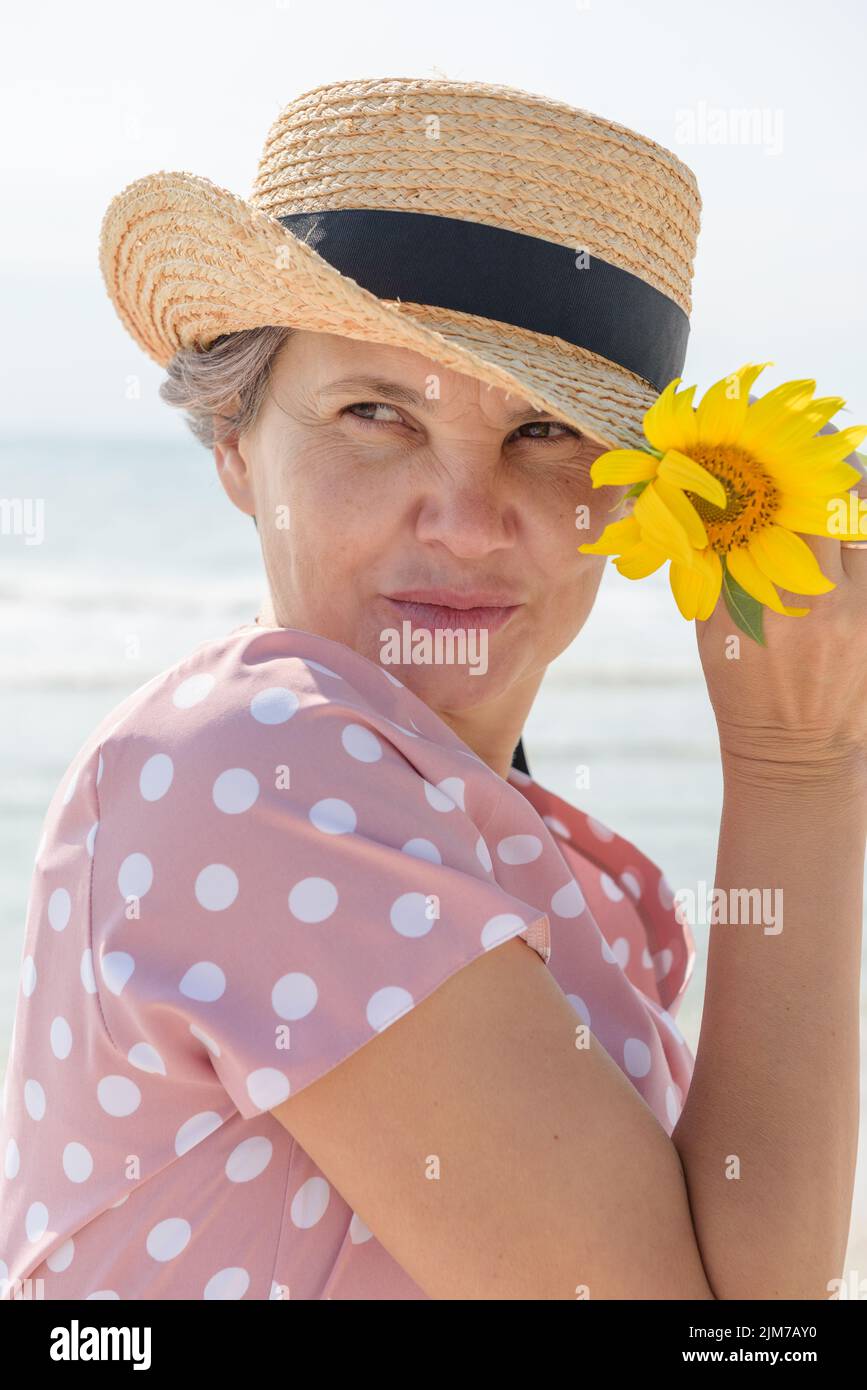 Frau in einem rosa Kleid mit weißen Tupfen, die Strohhut und Sonnenblume in der Hand halten und vor dem Hintergrund des Meeres wegschauen und scharfsinnig lächeln. Stockfoto