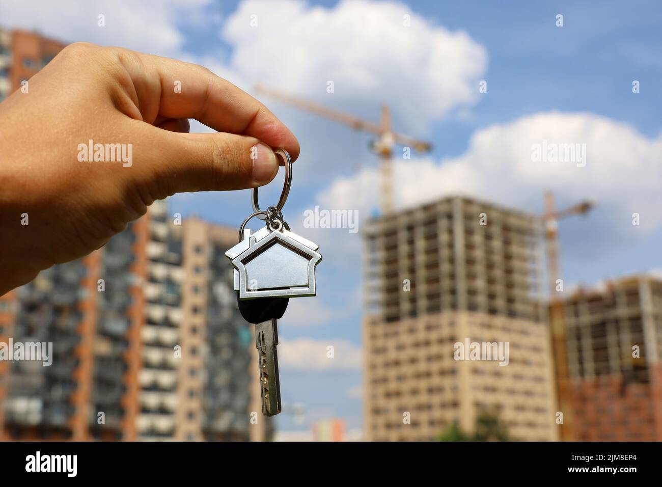 Immobilienmakler, Schlüsselanhänger in Form eines Hauses und Schlüssel in männlicher Hand auf dem Hintergrund von Baukräne und Neubauten. Byung Wohnung oder Miete Stockfoto