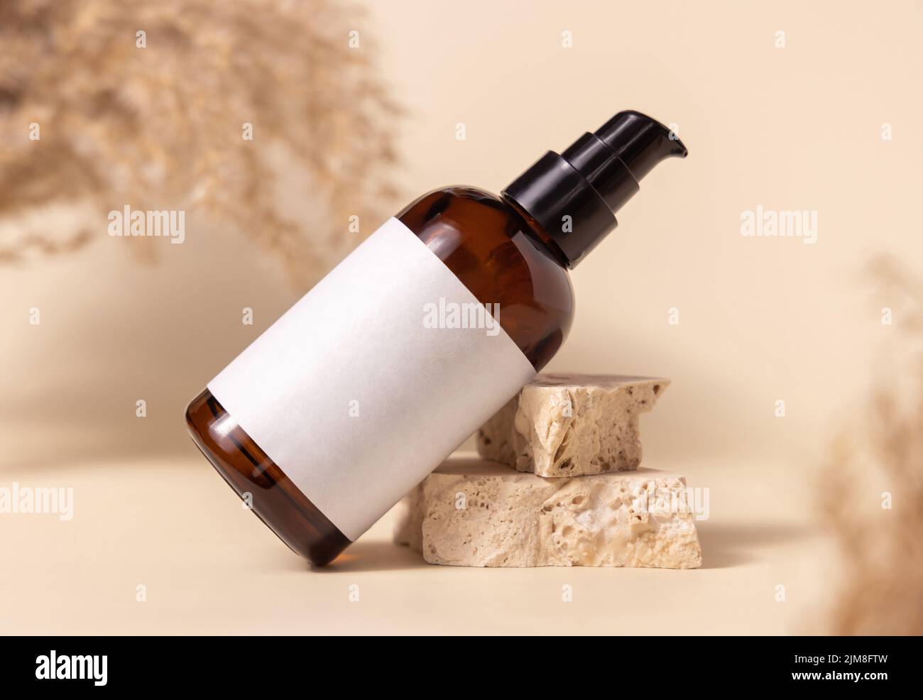 Braune Glas-Kosmetikflasche mit leerem Etikett auf Steinen in der Nähe von Pampas Gras auf hellgelb, Nahaufnahme, Mockup. Hautpflegeprodukt. Natürliche Öko-Frien Stockfoto