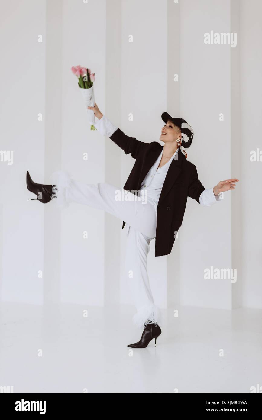 Eine fröhliche, stilvolle Frau in einem modischen Outfit, ein hellweißer Anzug mit Federn, eine schwarze Jacke, eine Baseballmütze und mit einem Blumenstrauß in ihrem h Stockfoto