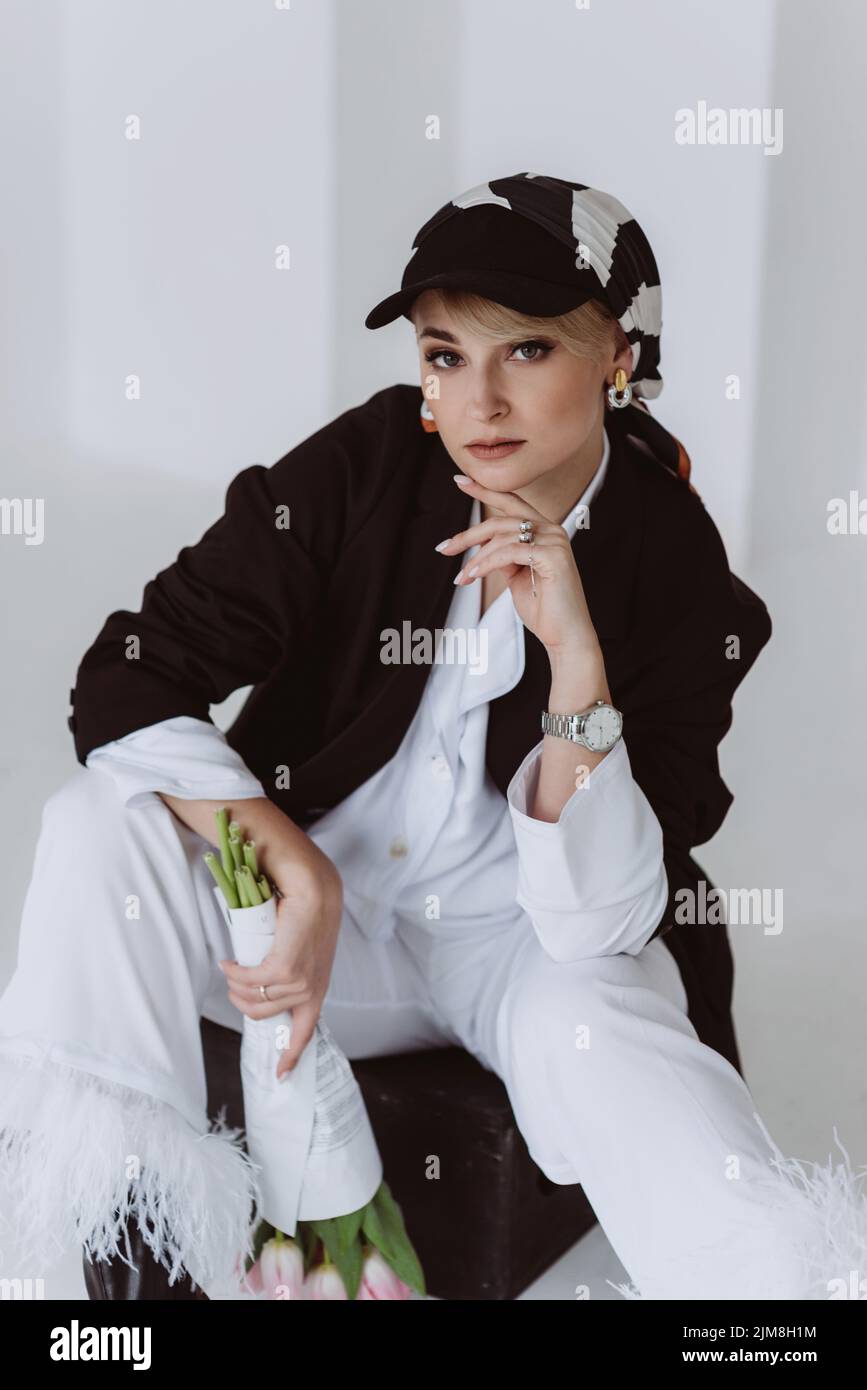 Eine wunderschöne alleinstehende Frau mittleren Alters in einem hellweißen Anzug und einer schwarzen Jacke mit einem Blumenstrauß posiert für die Kamera. Weicher, selektiver Fokus. Stockfoto