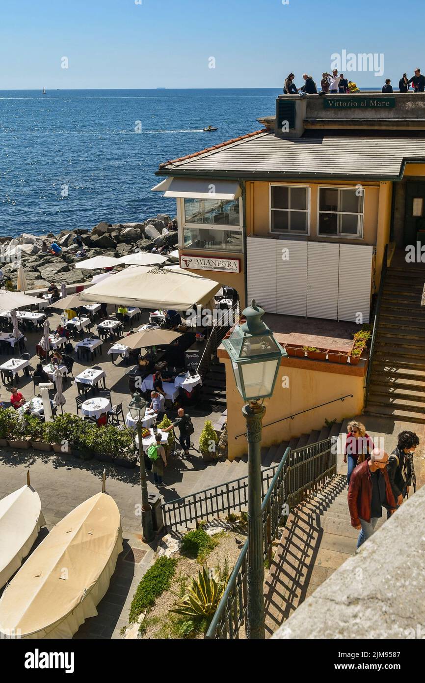 Blick auf die Uferpromenade des Fischerdorfes mit Touristen auf dem Aussichtspunkt und im Freien cafè am Ostermontag, Boccadasse, Genua, Italien Stockfoto