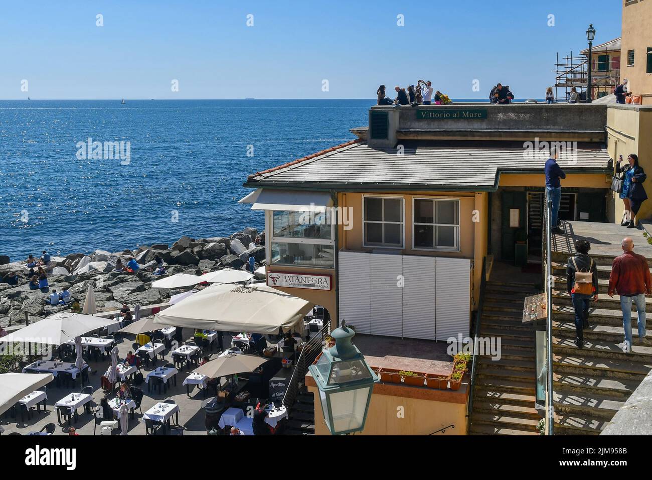 Blick auf die Uferpromenade des Fischerdorfes mit Touristen auf dem Aussichtspunkt und im Freien cafè am Ostermontag, Boccadasse, Genua, Italien Stockfoto