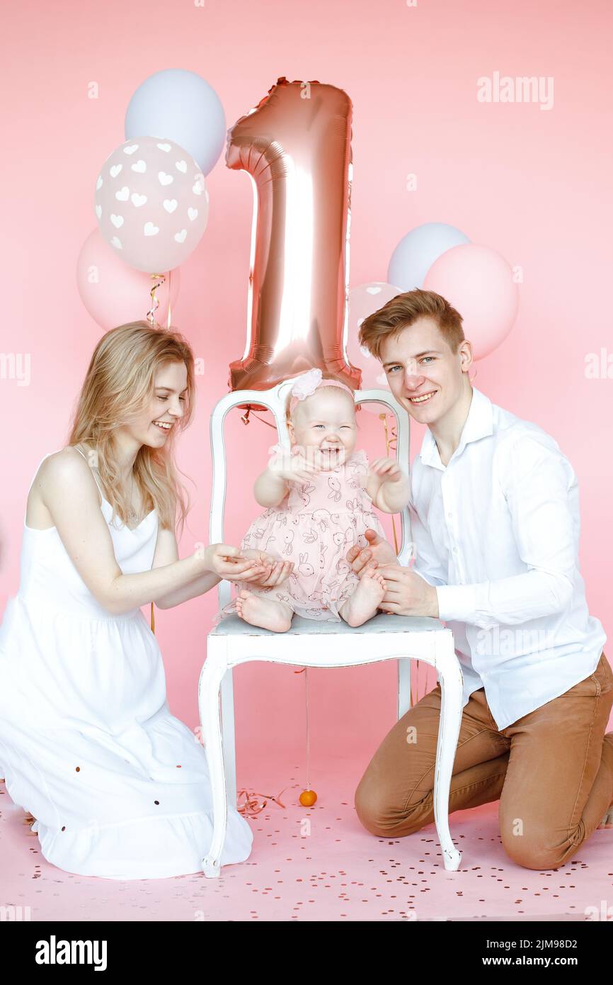 Vertikale lachende Familie von Mann, Frau und einem Jahr Kleinkind sitzen und feiern Geburtstag. Pinke Ballone und funkelt Stockfoto