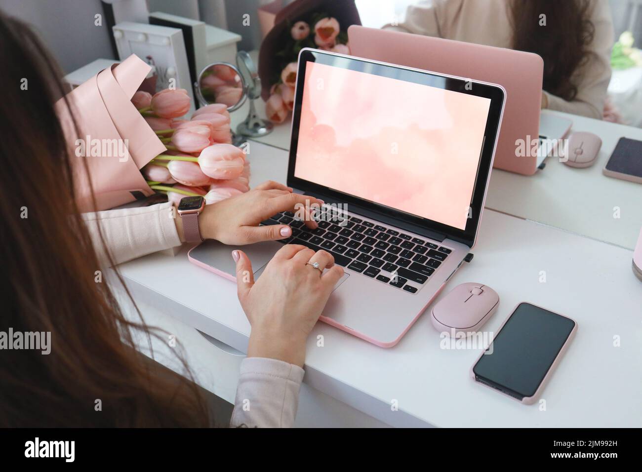 Weibliche Hände mit Laptop. Weibliche Büro Schreibtisch Arbeitsplatz Homeoffice Nachbau mit Laptop, rosa Tulpenblüten Blumenstrauß, Smartphone und rosa Accessoires. Stockfoto