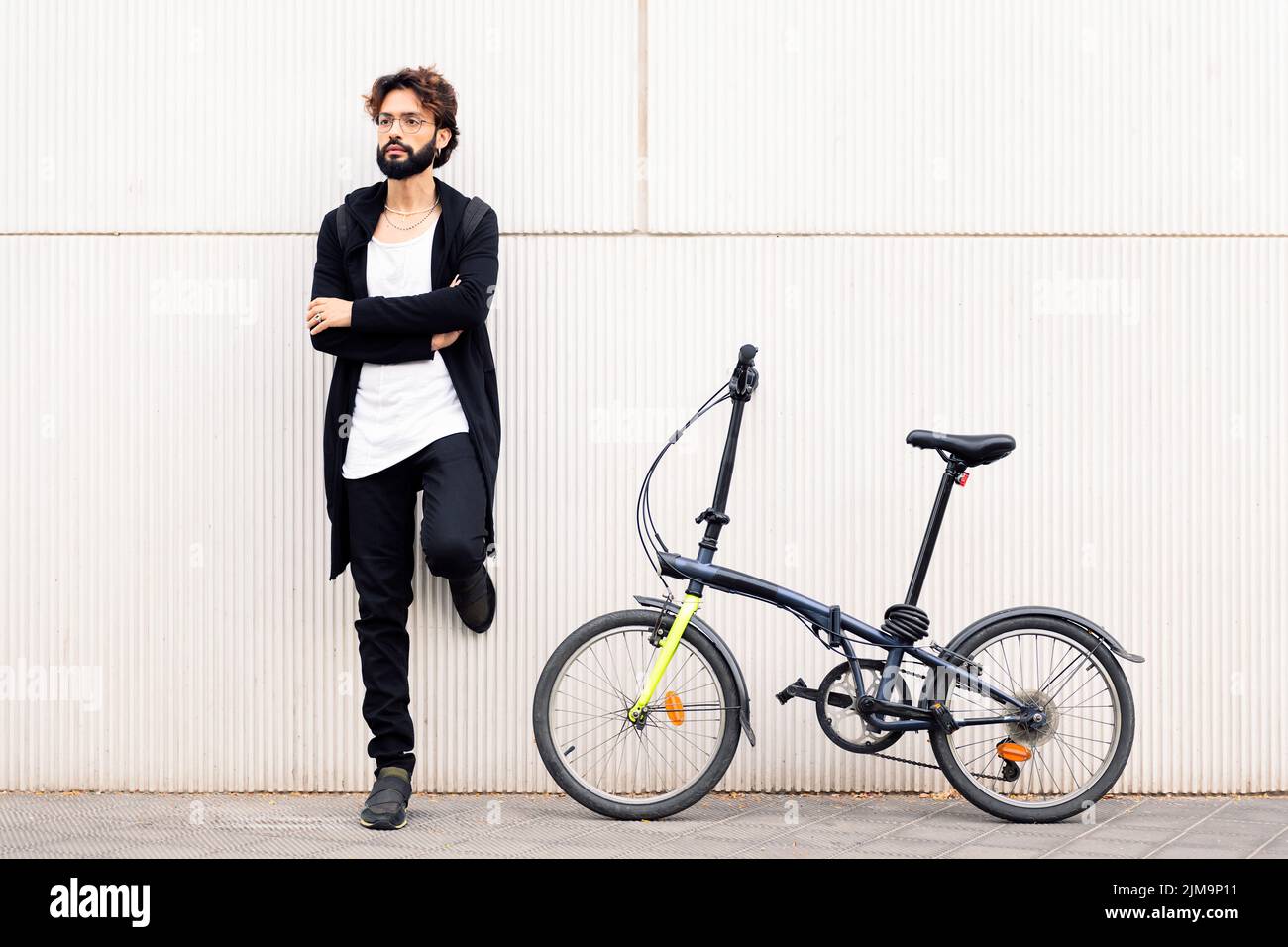 Stilvoller junger Mann mit Bart an einer weißen Wand neben seinem Faltrad gelehnt, Konzept von urbanem Lifestyle und nachhaltiger Mobilität, Kopierraum fo Stockfoto