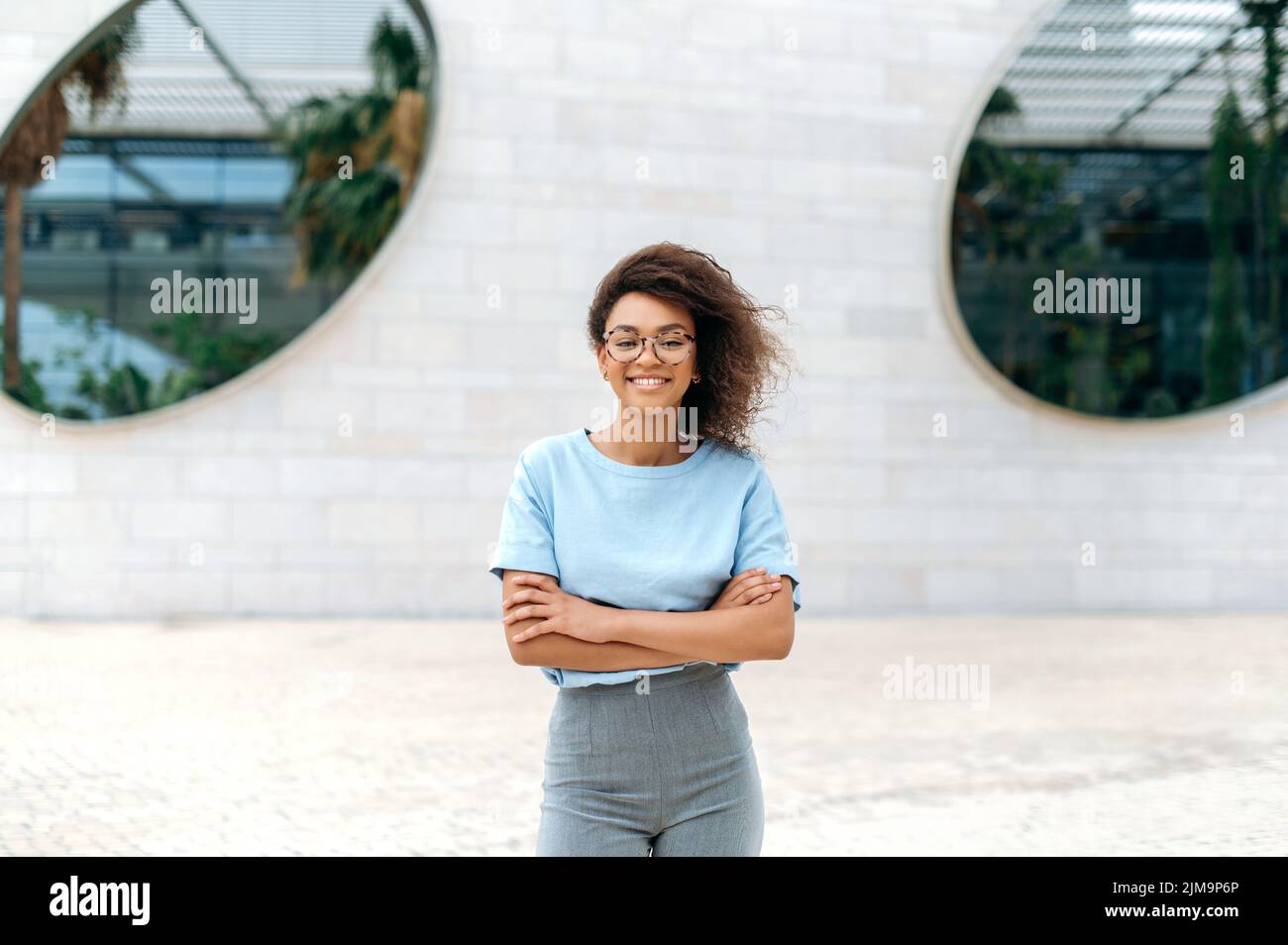 Porträt einer positiv eleganten, hübschen afroamerikanischen jungen Frau mit lockigen Haaren, Brille, blauem Hemd tragend, mit gekreuzten Armen im Freien stehend, lächelnd auf die Kamera schauend Stockfoto