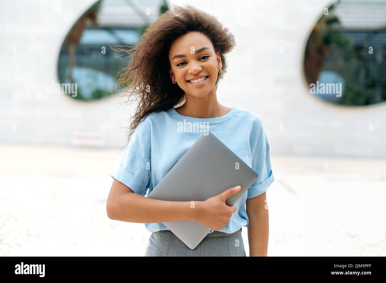 Elegante afroamerikanische junge Frau mit lockigen Haaren, freiberuflich tätige weibliche Mixed-Race-Mitarbeiterin, Studentin, mit Brille, in einem blauen Hemd, Laptop haltend, im Freien stehend, Kamera anblickend, glücklich lächelnd Stockfoto
