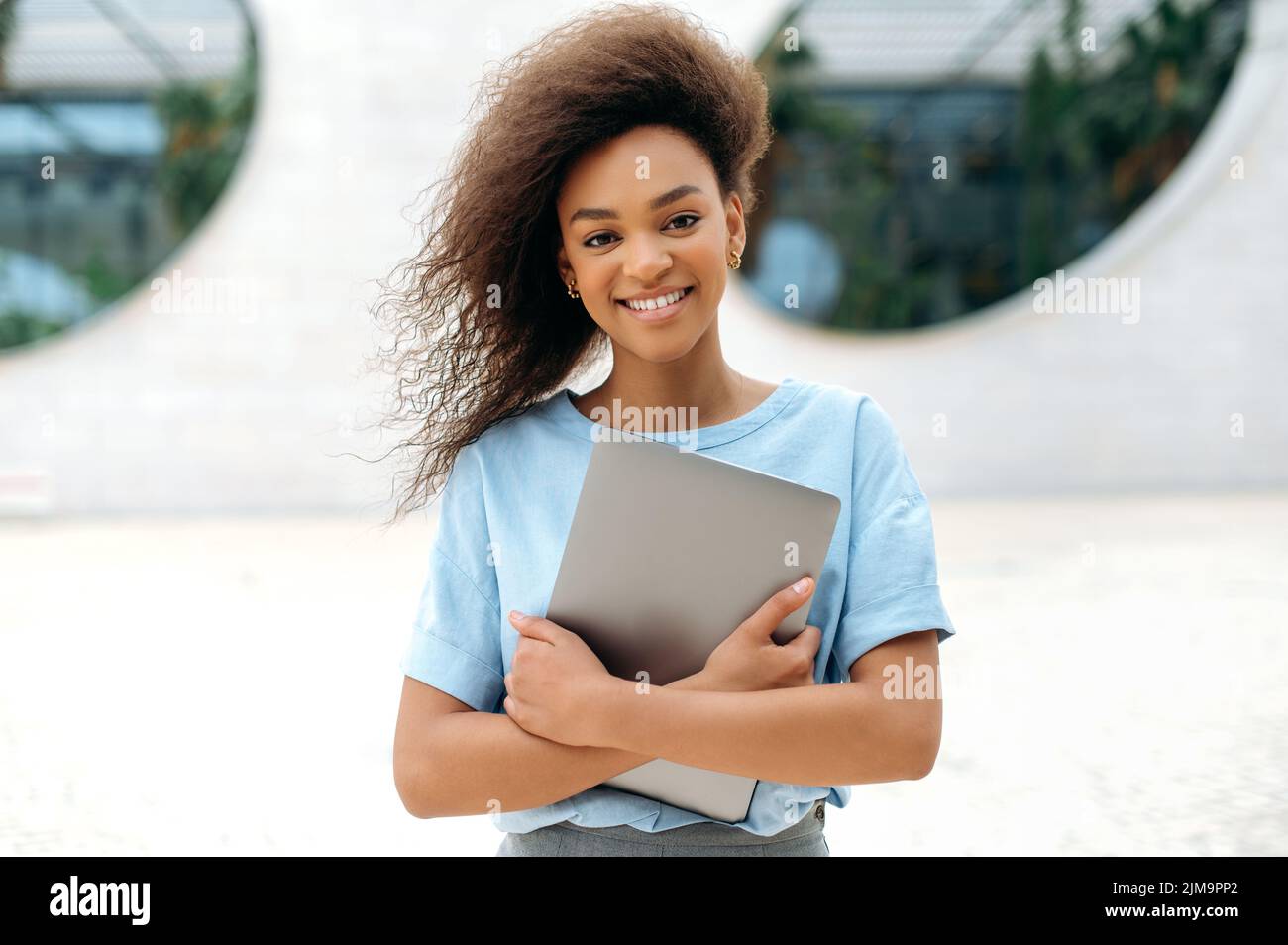 Freundliche, positive, lockige afroamerikanische junge Geschäftsfrau, Freiberuflerin, Studentin, mit Brille, in einem blauen Hemd, Laptop haltend, im Freien stehend, Kamera anblickend, glücklich lächelnd Stockfoto