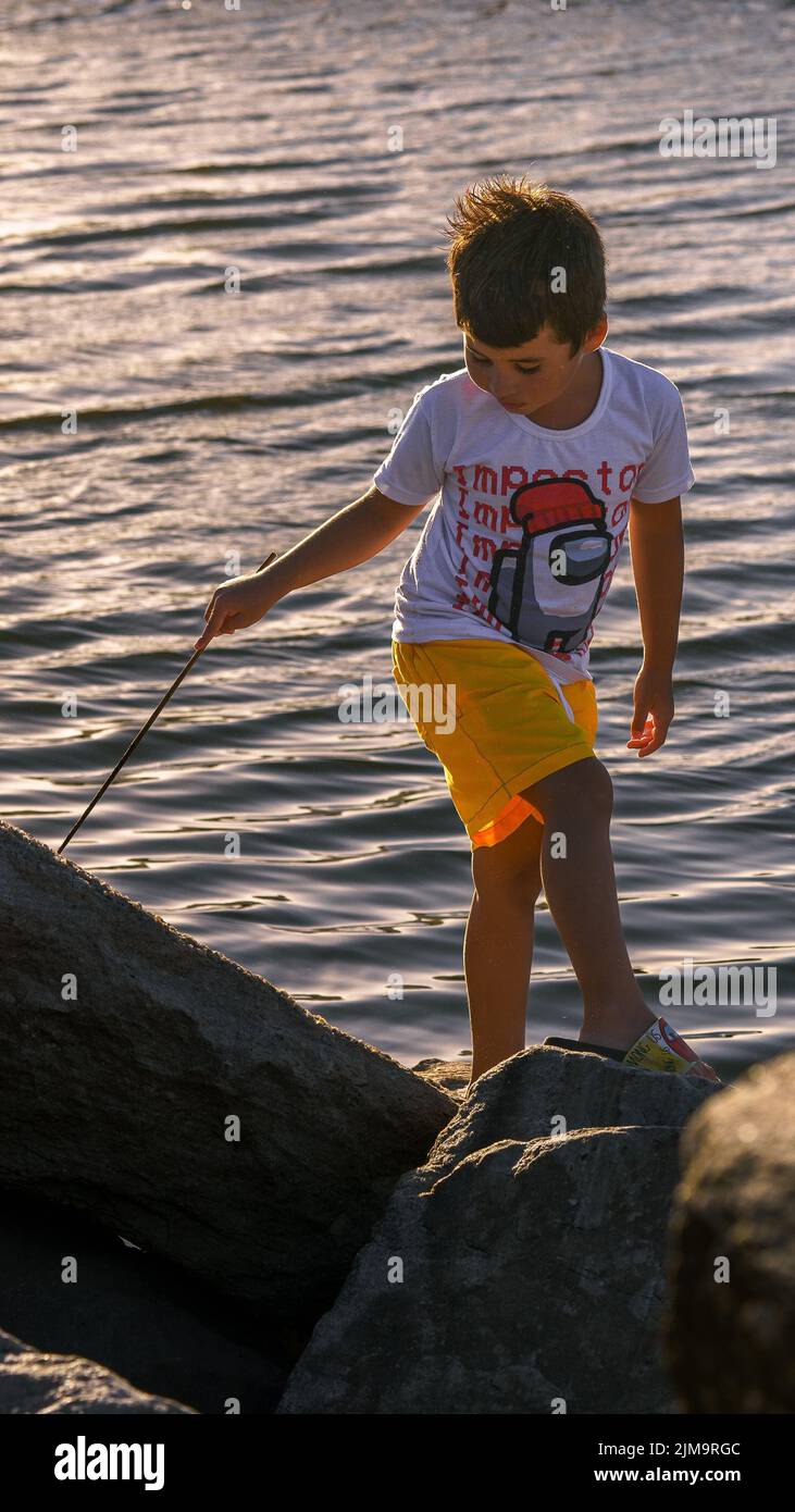 Eine vertikale Aufnahme eines Kindes auf den Felsen, das einen Holzstock am Fluss hält Stockfoto