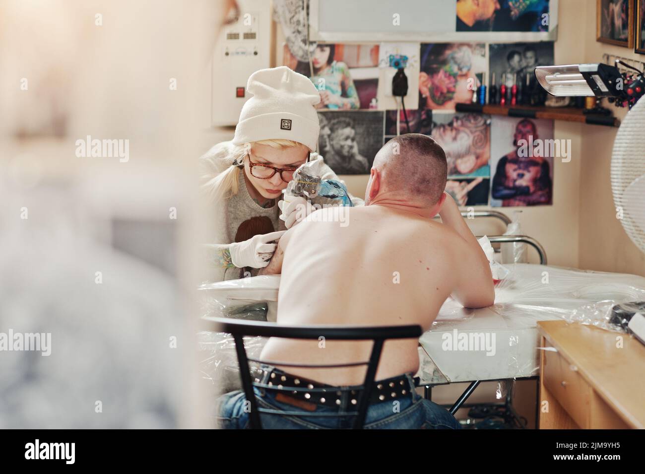 Junge blonde Frau Meister Tätowierer in Gläser und Hut mit Tattoo-Maschine von Hand machen tattoo-Dreamcatcher zur hand des Menschen. Stockfoto