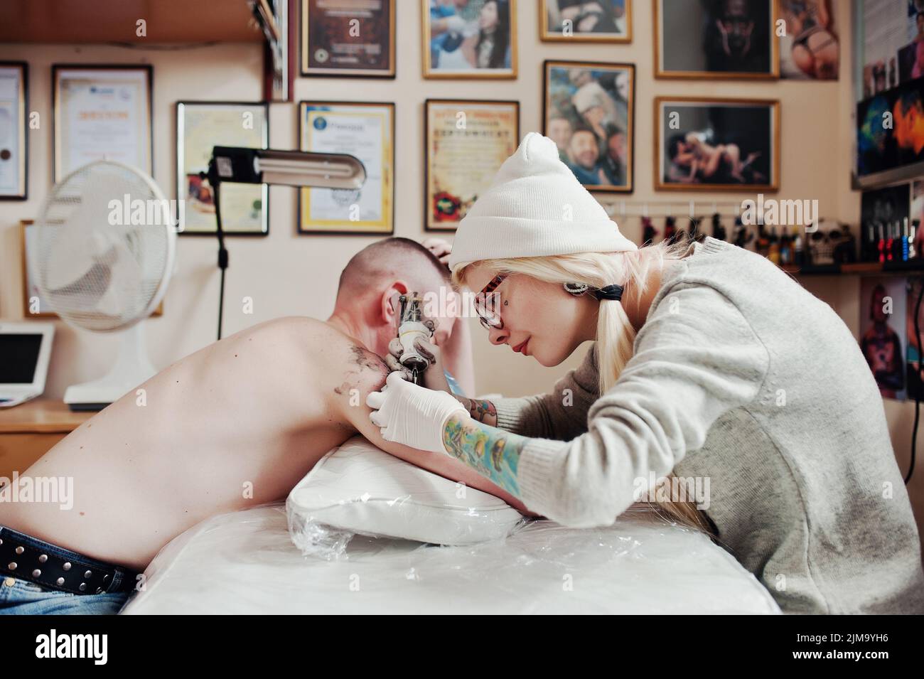 Junge blonde Frau Meister Tätowierer in Gläser und Hut mit Tattoo-Maschine von Hand machen tattoo-Dreamcatcher zur hand des Menschen. Stockfoto