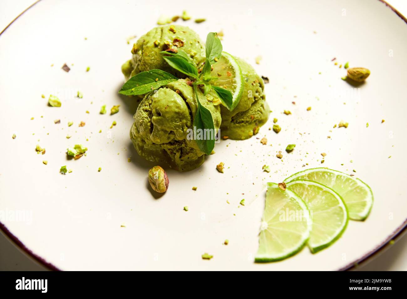 Selektive Konzentration auf Pistazien-Sorbet-Eiscreme mit bestreuten Nüssen und grünem Matcha-Pulver auf weißem Teller, garniert mit frischem, saftigem Limettenschrot Stockfoto