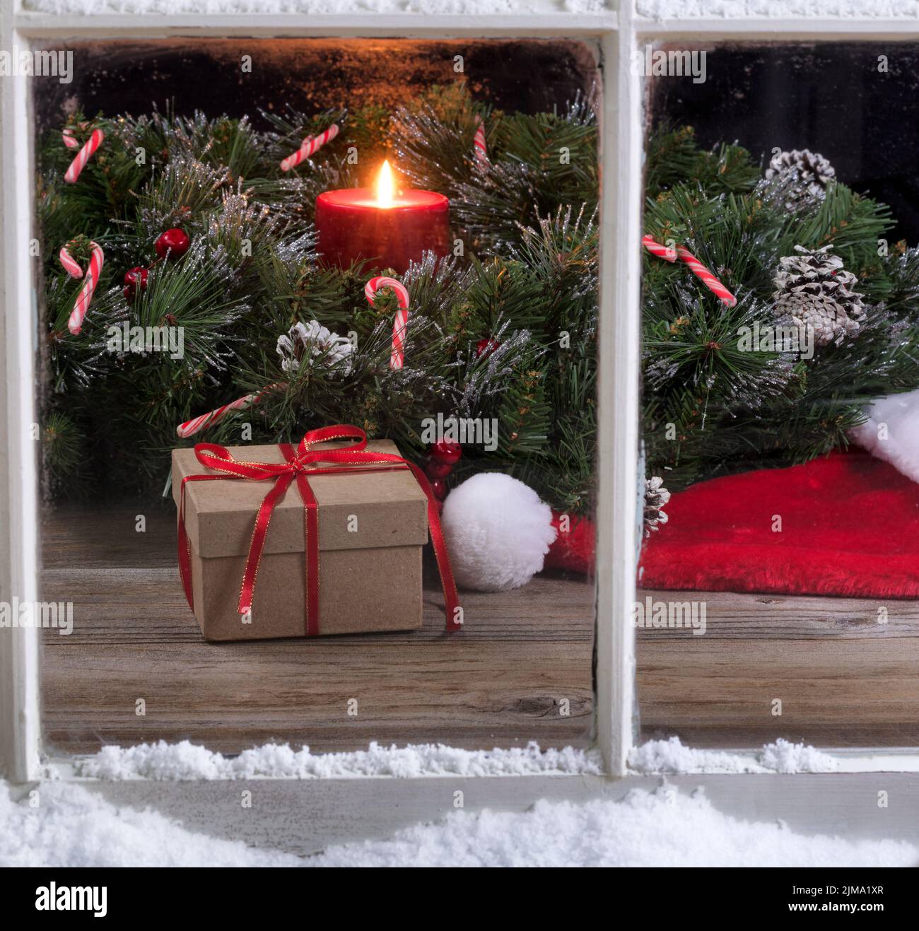 Verschneite Fensteransicht der Geschenkbox und der Weihnachtsmütze mit brennender Kerze im Hintergrund Stockfoto Verschneite Fensteransicht der Geschenkbox und der Weihnachtsmütze mit brennender Kerze im Hintergrund Stockfoto