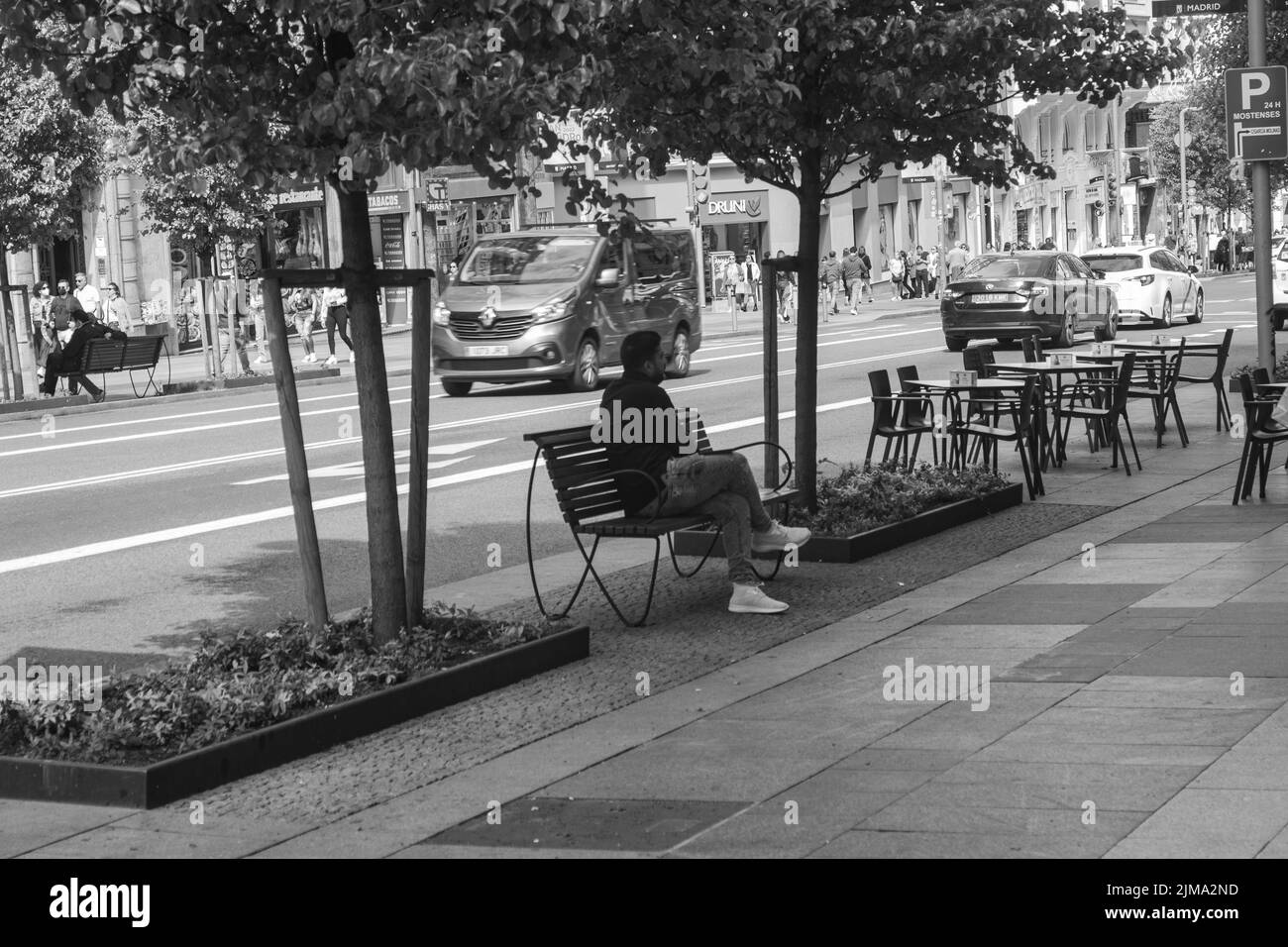 Eine Person, die auf einer Bank in der Straße in Madrid sitzt und den Rest in Graustufen schlendert Stockfoto