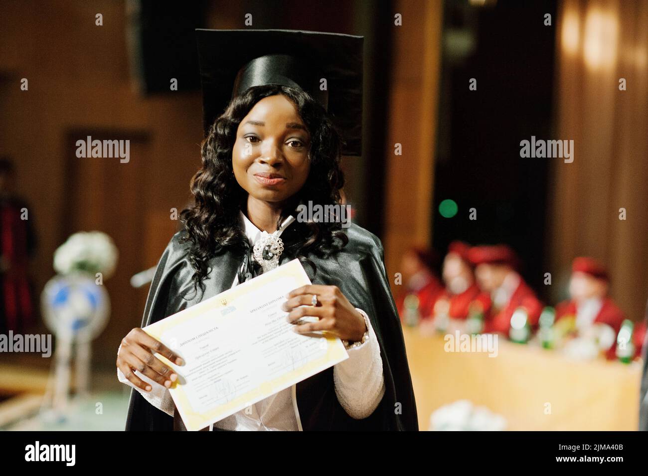 Glückliches schönes schwarze afroamerikanische Mädchen mit Hut und Mantel Absolventen bei Zeremonie Studium mit Diplom bei der hand Stockfoto