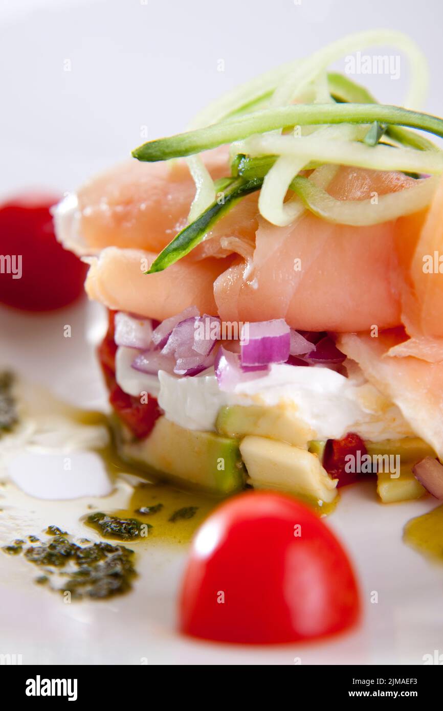 Gourmet-Teller geräucherten Lachs auf Käse, Avocado, rote Zwiebel und beträufelt mit Basilikum-Pesto, garniert mit Gurken-Streifen. Stockfoto