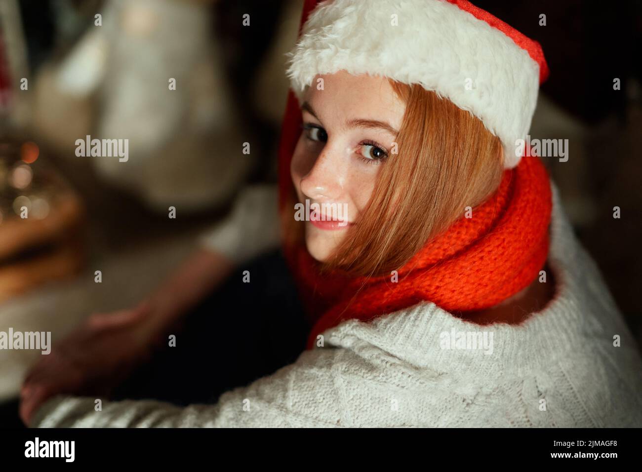 Fröhliche kaukasische weibliche Teenager in Weihnachtsmannhut und rotem Schal mit Neujahrsdekoration und schwachem Licht. Junges Mädchen feiert Neujahr 2023. Hochwertige Fotos Stockfoto