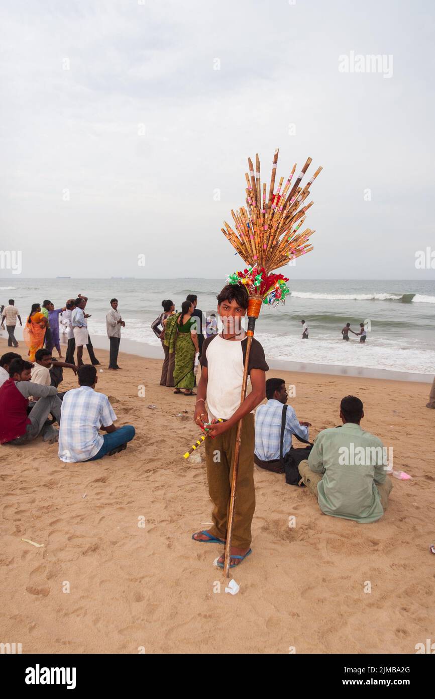 Ein einheimischer Junge, der Spielzeug und Souvenirs verkauft, posiert für ein Foto am Marina Beach in Chennai, während andere Leute sich im Rücken Vergnügen Stockfoto