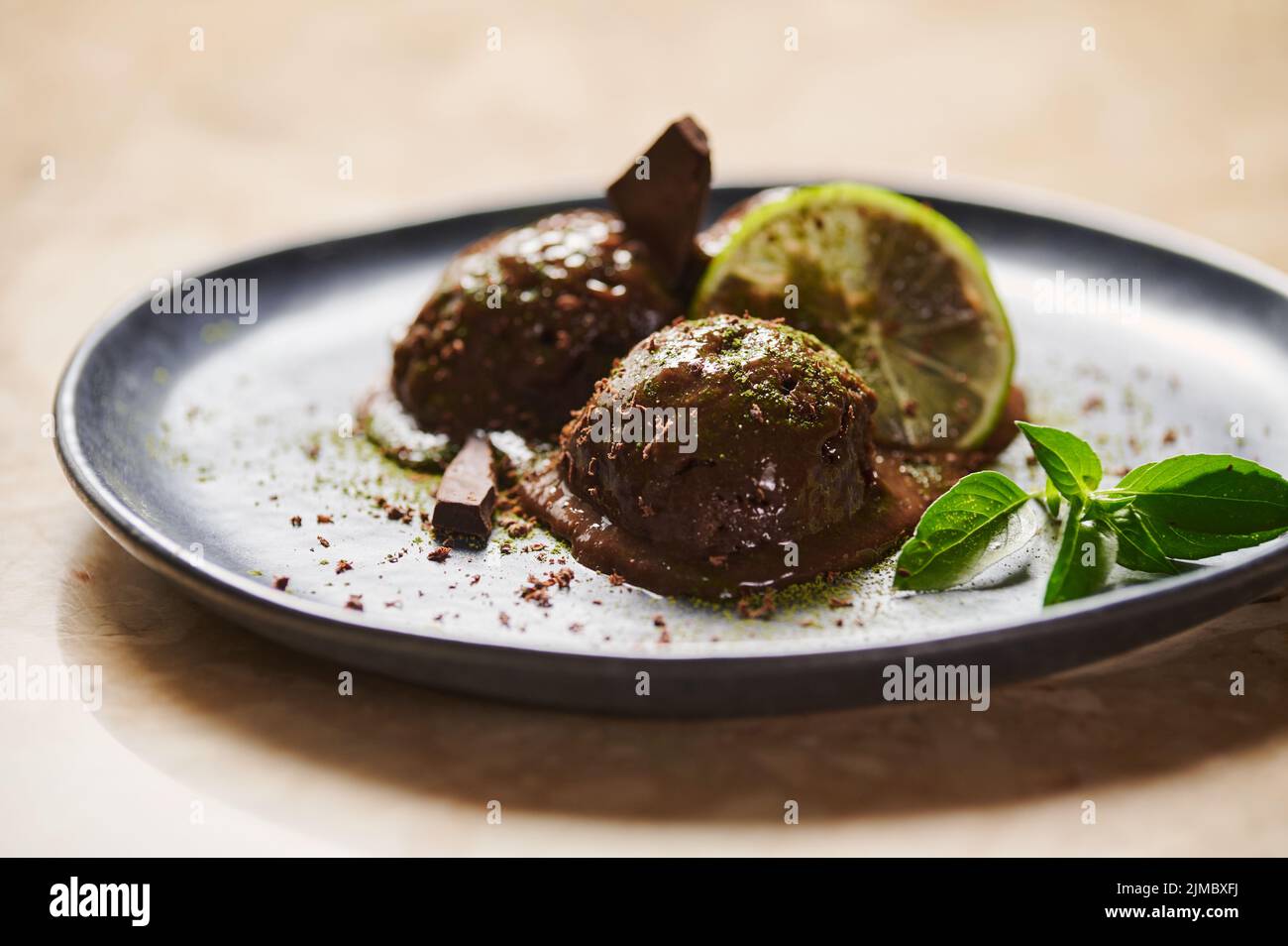 Kugeln Schokoladensorbet, mit bestreutem Matcha-Pulver, Limettenscheibe, Zitronenbasil auf dunkelblauem Teller. Selektiver Fokus Stockfoto