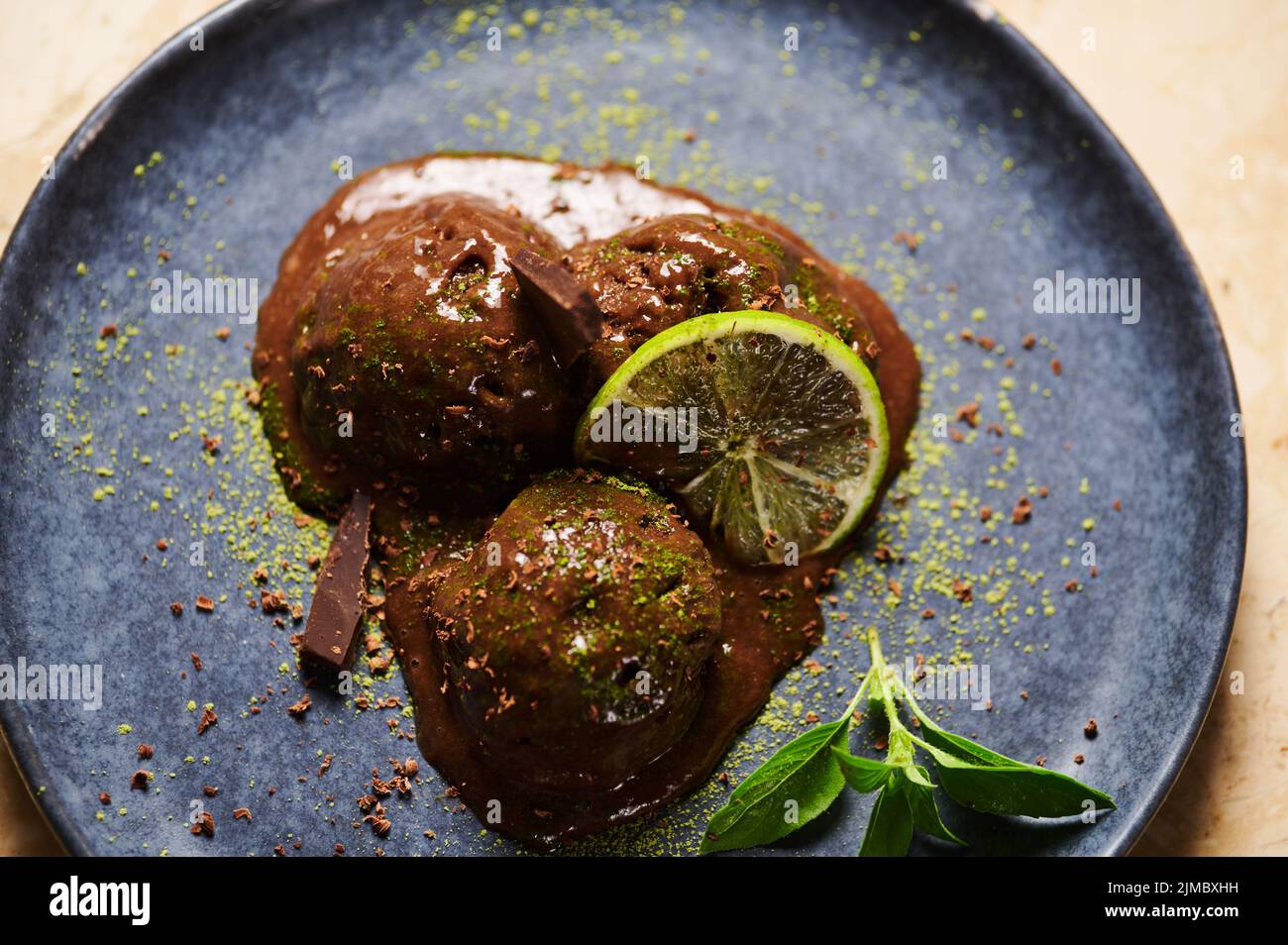 Kugeln Schokoladensorbet, mit bestreutem grünem Matcha-Pulver, Limettenscheibe, Zitronenbasil auf dunkelblauem Teller. Flach liegend Stockfoto