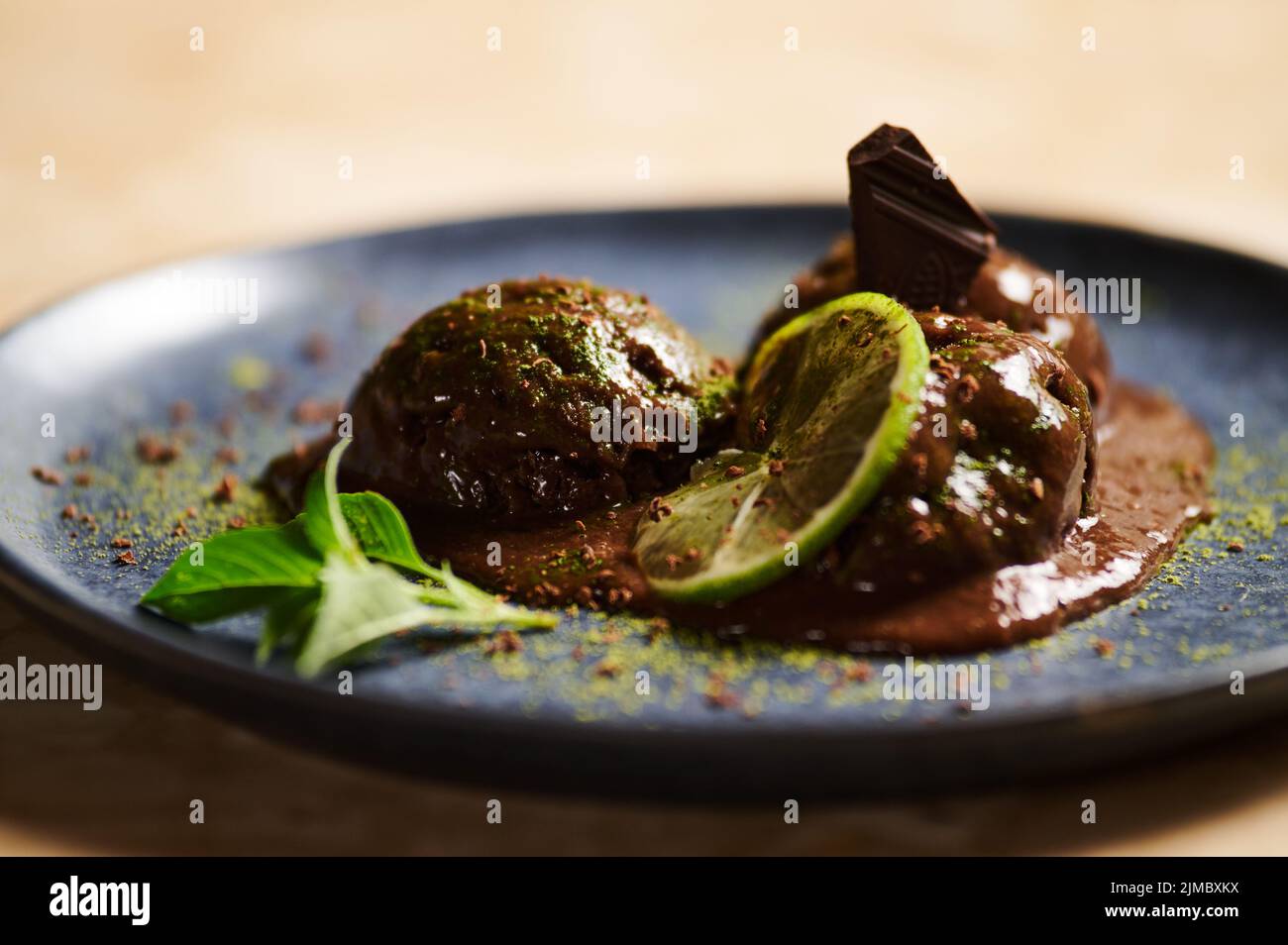 Kugeln Schokoladensorbet, mit bestreutem Matcha-Pulver, Limettenscheibe, Zitronenbasil auf dunkelblauem Teller. Selektiver Fokus Stockfoto