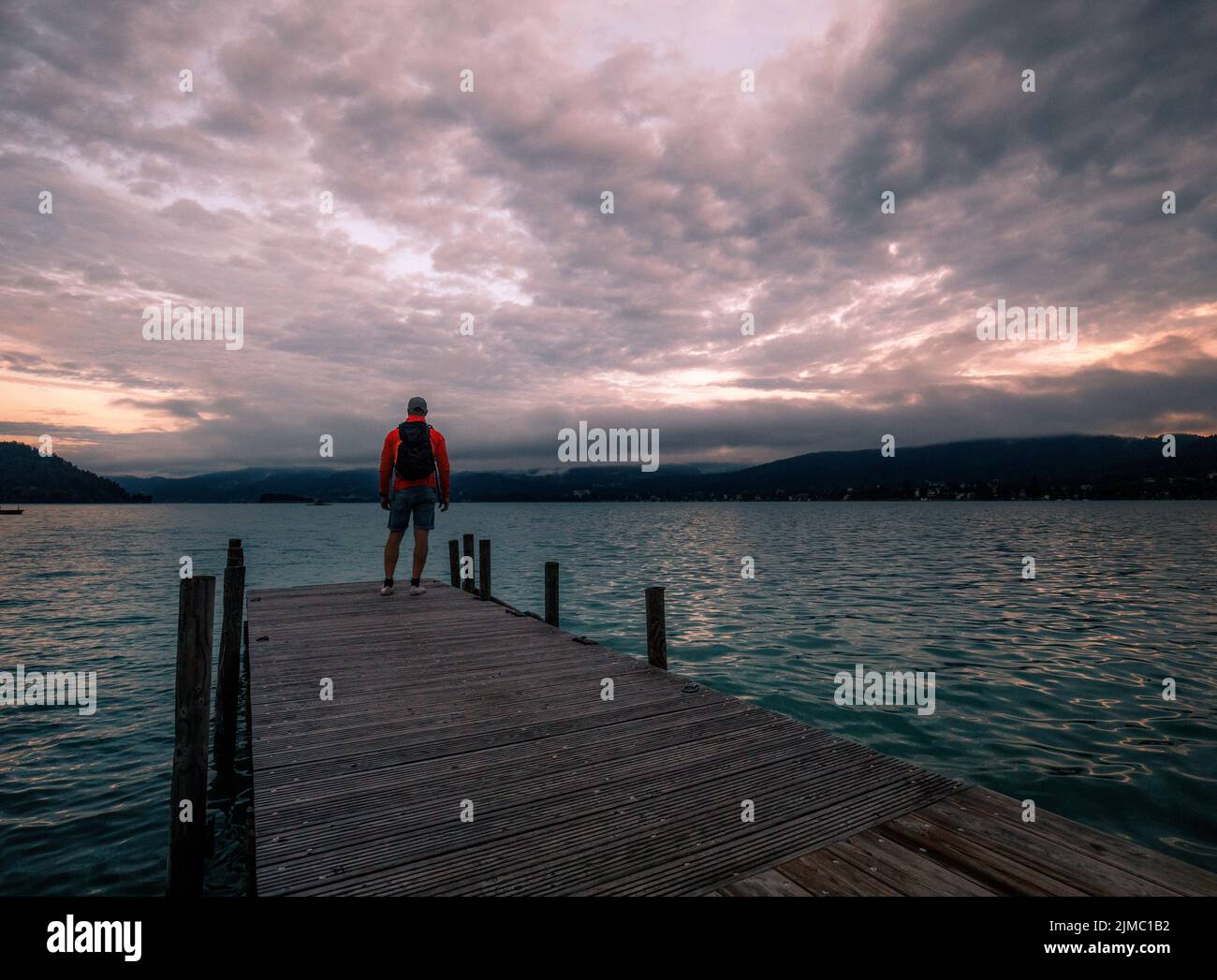 Einsamer Mann, der am Rand des hölzernen Piers steht und auf den ruhigen See und den Wald auf der anderen Seite blickt Stockfoto