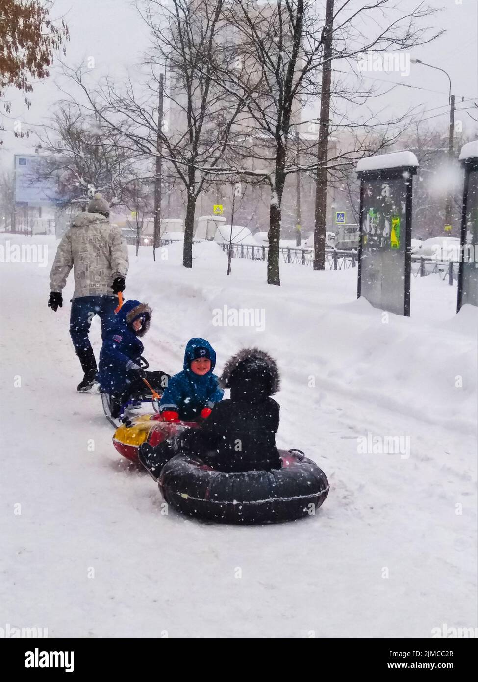 Ein Vater trägt seine drei Kinder auf einem Dampfzug aus Schlitten und zwei Airbags in einem Schneesturm entlang einer verschneiten Winterstraße. Stockfoto