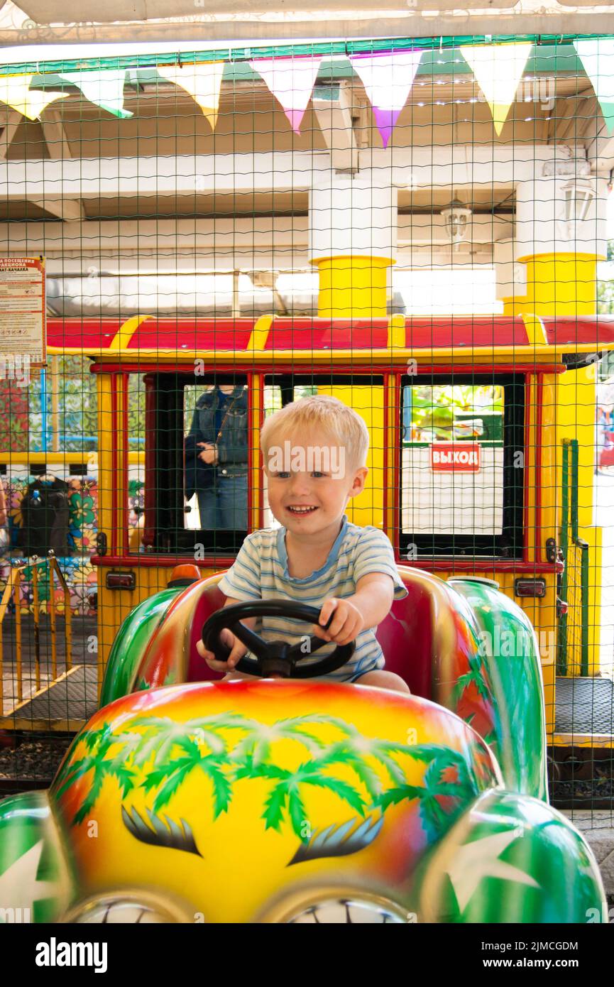 Ein sehr fröhlicher und glücklicher kleiner blonder Junge von drei Jahren fährt im Sommer an der Riviera Amu in einem gelben Auto mit einem grünen Auto Stockfoto