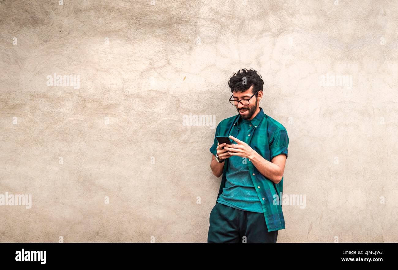 Glücklicher gutaussehender Kerl, der sich mit seinem Handy an die Wand lehnte, glücklicher lateinischer Mann, der sein Handy an einer Wand mit Platz für Kopien lehnte, Person Stockfoto