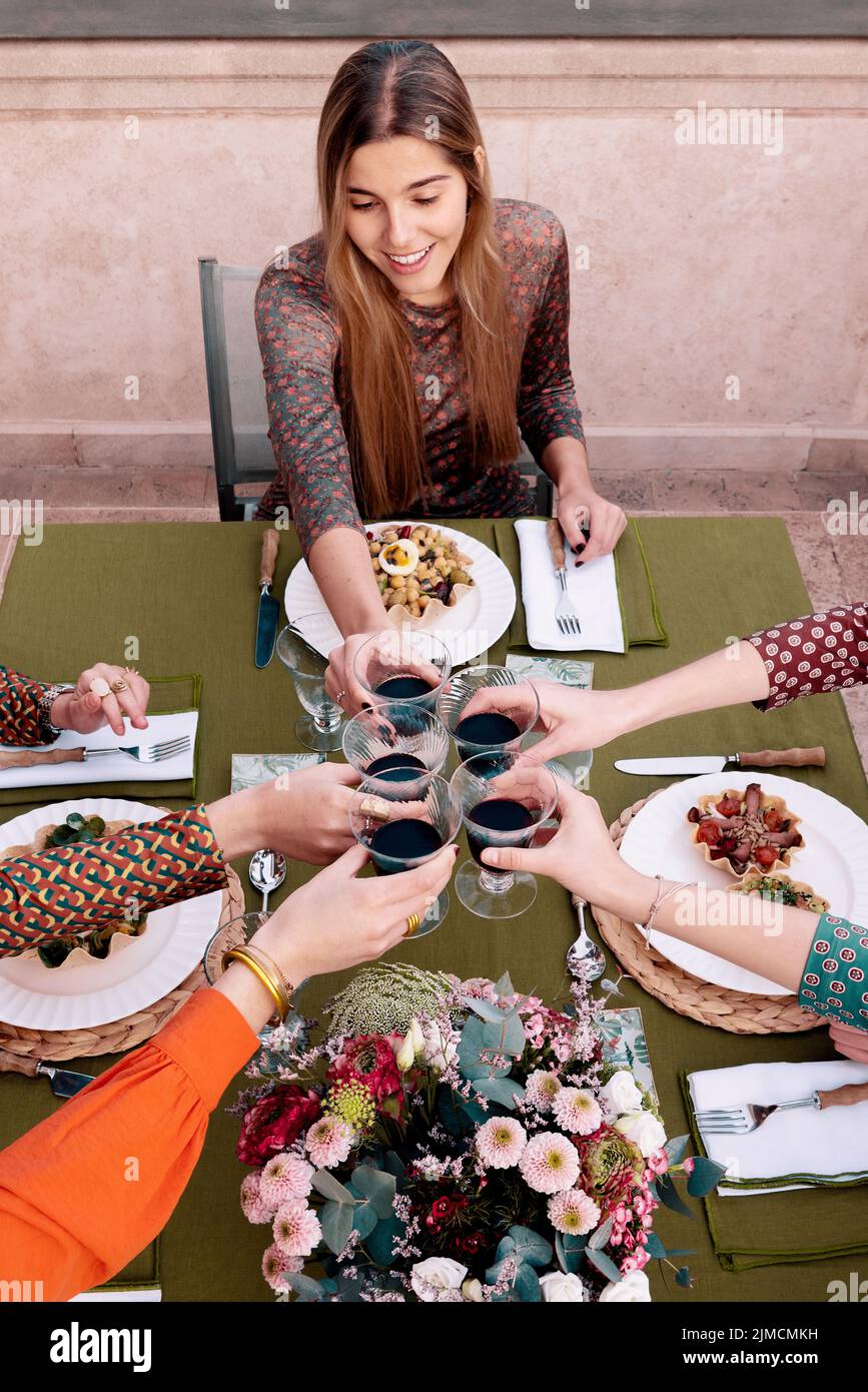 Gruppe positiver Freundinnen, die mit Weinbässen toasten, während sie am Tisch mit Salaten auf der Terrasse essen Stockfoto