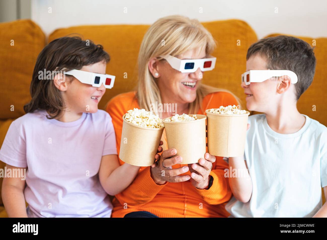 Positive Mutter und Kinder in 3D Gläsern, die leckeres Popcorn halten und sich gegenseitig angucken, während sie sich auf der Couch im Wohnzimmer einen Film ansehen Stockfoto