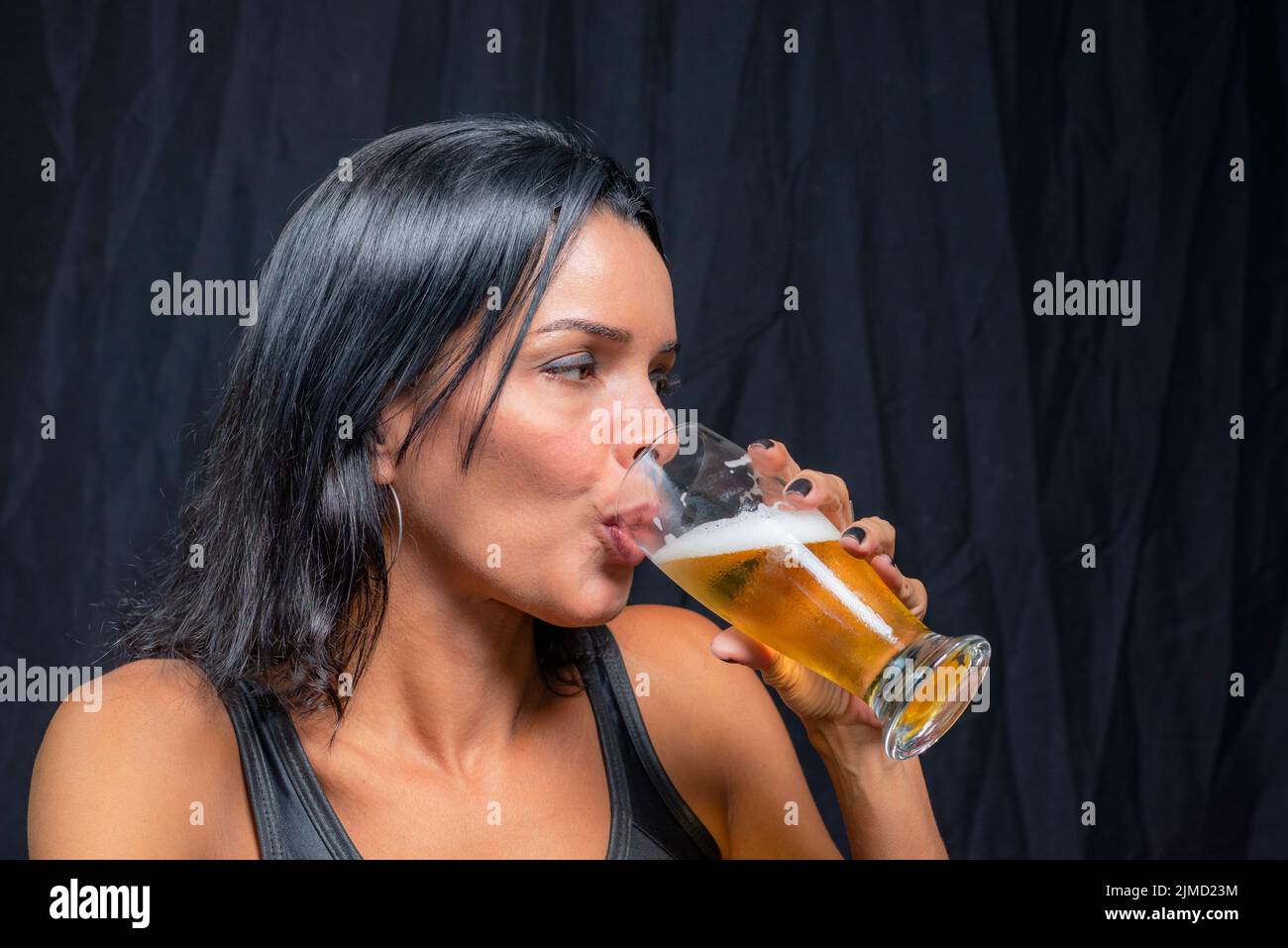 Porträt einer jungen Frau im schwarzen T-Shirt, die Bier aus einem Glas trinkt. Vor schwarzem Studiohintergrund. Salvador, Brasilien. Stockfoto