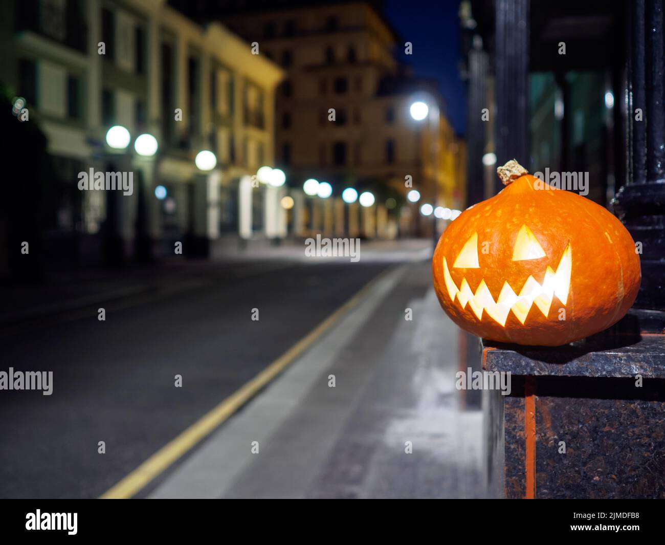 Halloween Kürbis auf einer verlassenen Stadtstraße in der Nacht. Die Lichter der Stadt und die alten Hochhäuser sind in verschwommenen Farben. Stockfoto