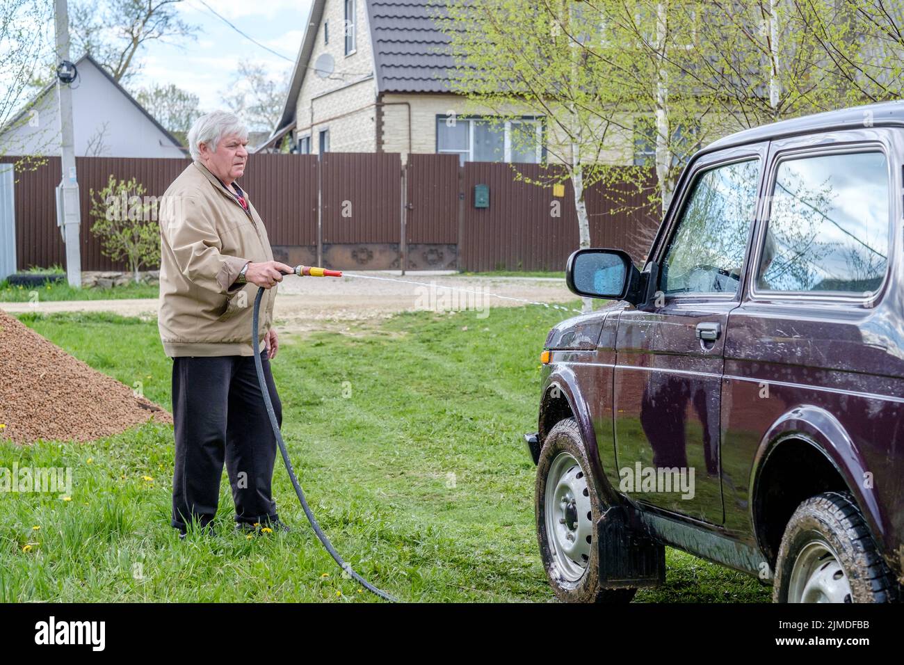 Ein grauhaariger älterer Mann wäscht ein Auto mit einem Gartenschlauch im Freien. Stockfoto