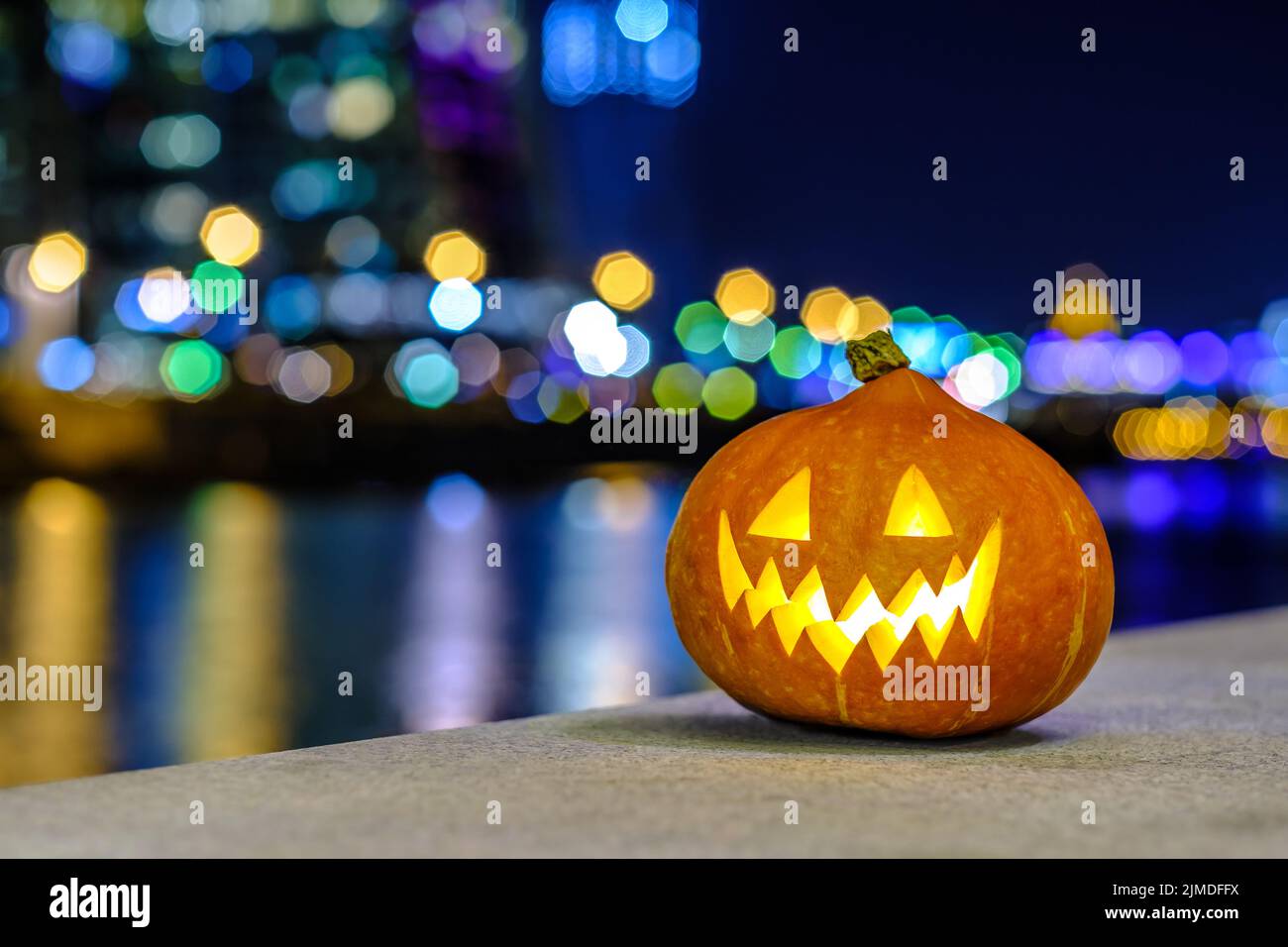 Halloween Kürbis auf dem Hintergrund der Nachtstadt. Verschwommene farbige Lichter werden im Wasser reflektiert. Nächtliches Stadtdekor mit einem Stockfoto