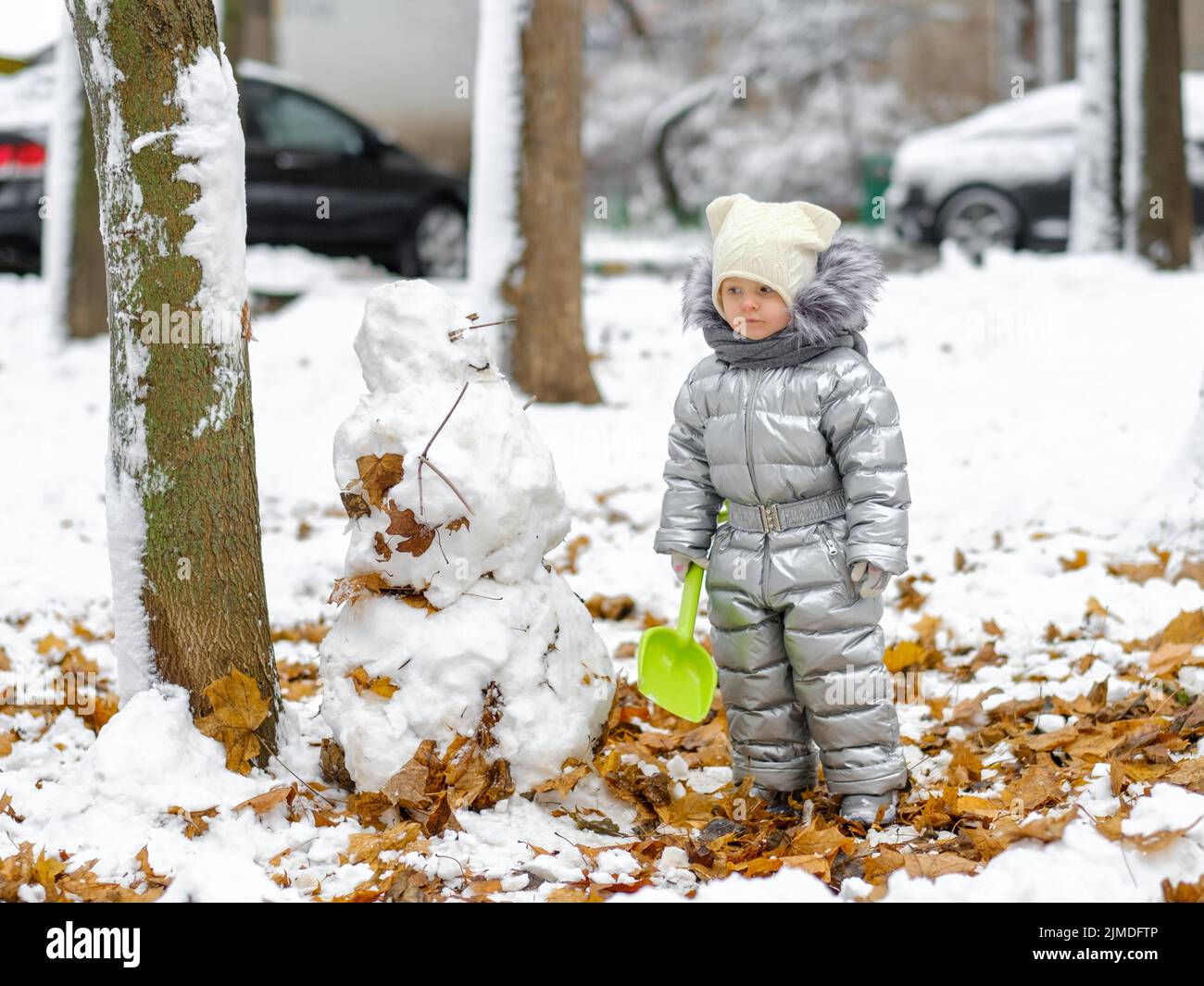 Ein lustiges Mädchen in einem silbernen Jumpsuit hält eine Spielzeugschaufel und modelliert einen Schneemann. Stockfoto