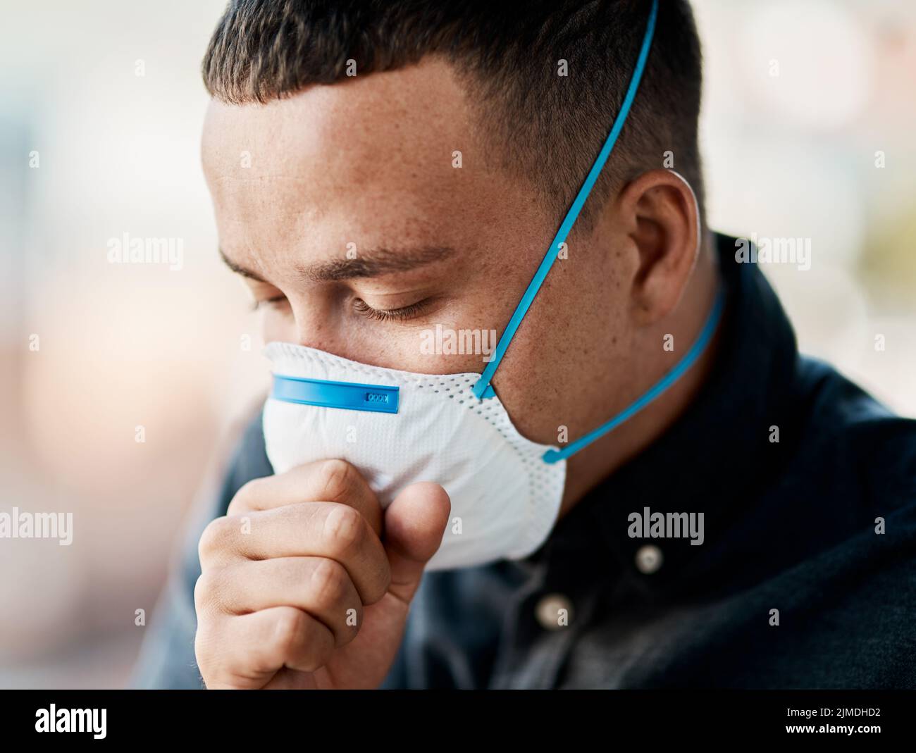 Heute ist es so viel mehr als nur ein Husten. Ein junger Mann hustet und trägt eine Maske vor einem Stadthintergrund. Stockfoto