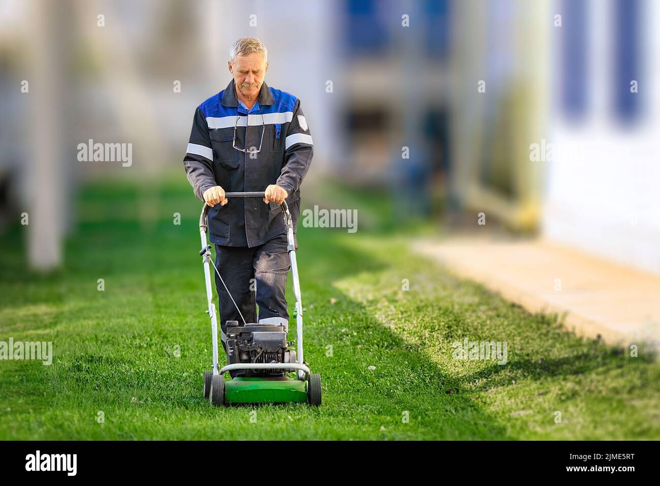 Ein älterer Arbeiter mäht an einem sonnigen Tag den Rasen. Ein Mann mit Arbeitskleidung arbeitet mit einem Rasenmäher auf einer grünen Wiese. Stockfoto
