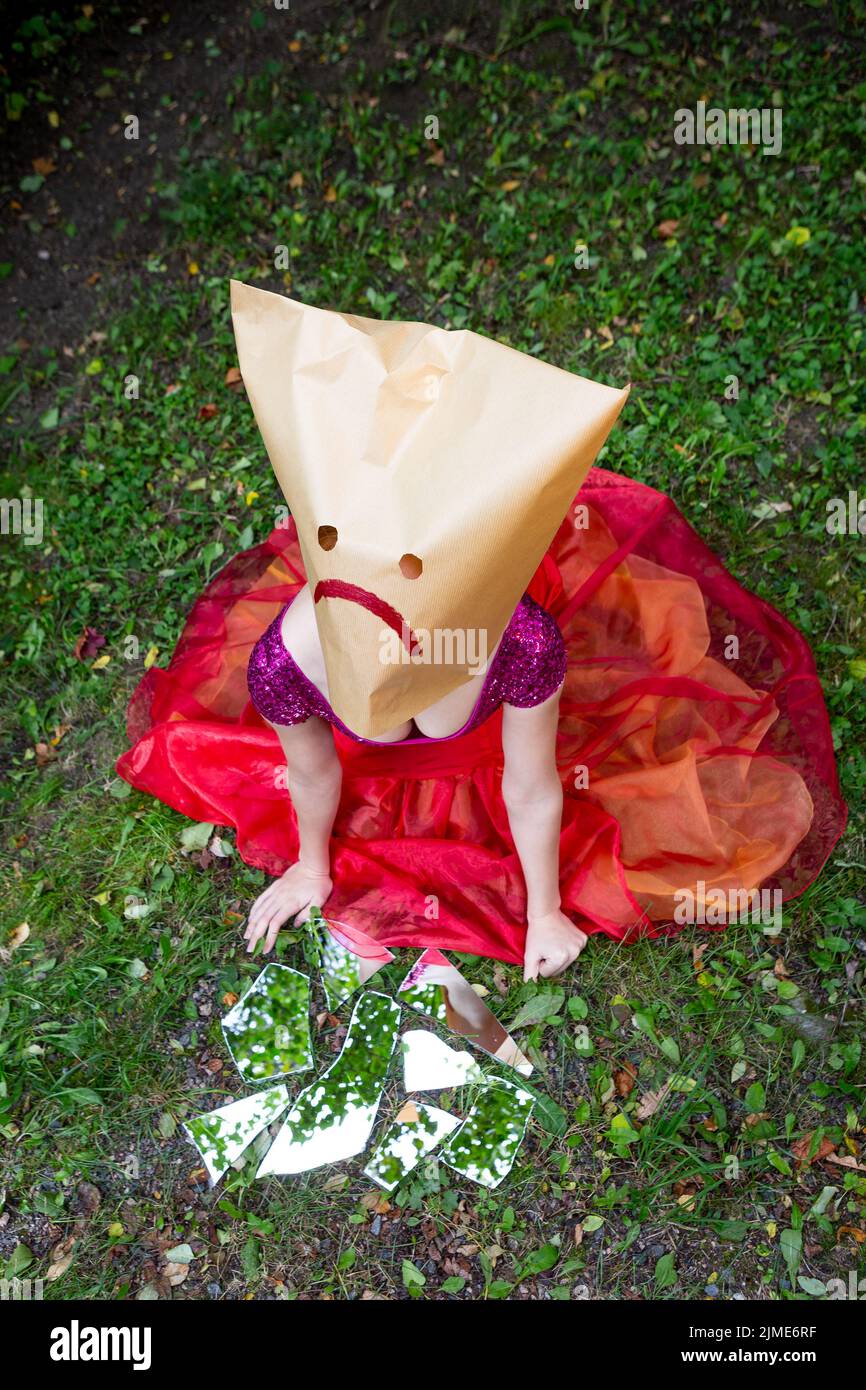 Eine junge Frau ohne Gesicht liegt auf den Knien auf dem Boden. Eine Frau schaut mit einem Papiersack auf ihrem Kopf und einem traurigen Gesicht darauf. Stockfoto