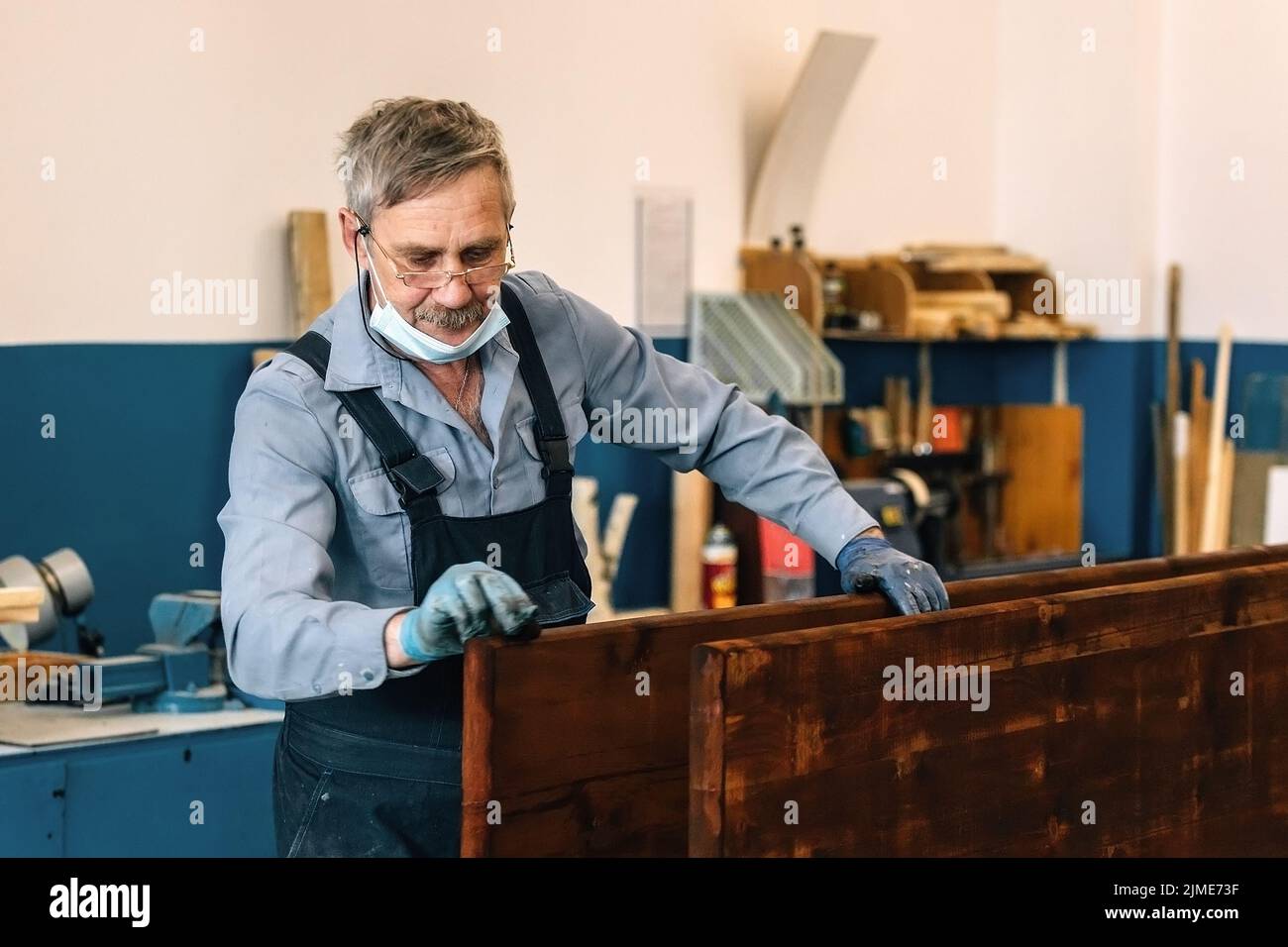 Ein älterer Mann malt in einer Schreinerei Holzbretter mit dunklem Lack. Ein Rentner in einer medizinischen Maske Stockfoto